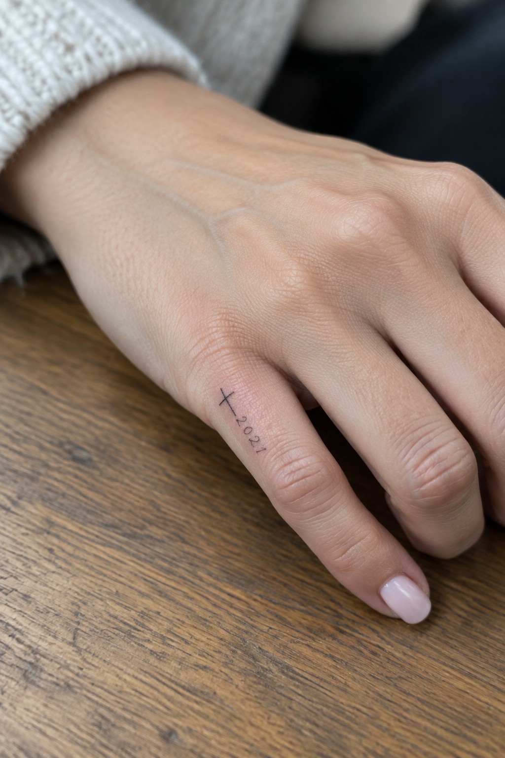 Hand with cross and 2021 tattoo on finger, pink nails, on wooden table