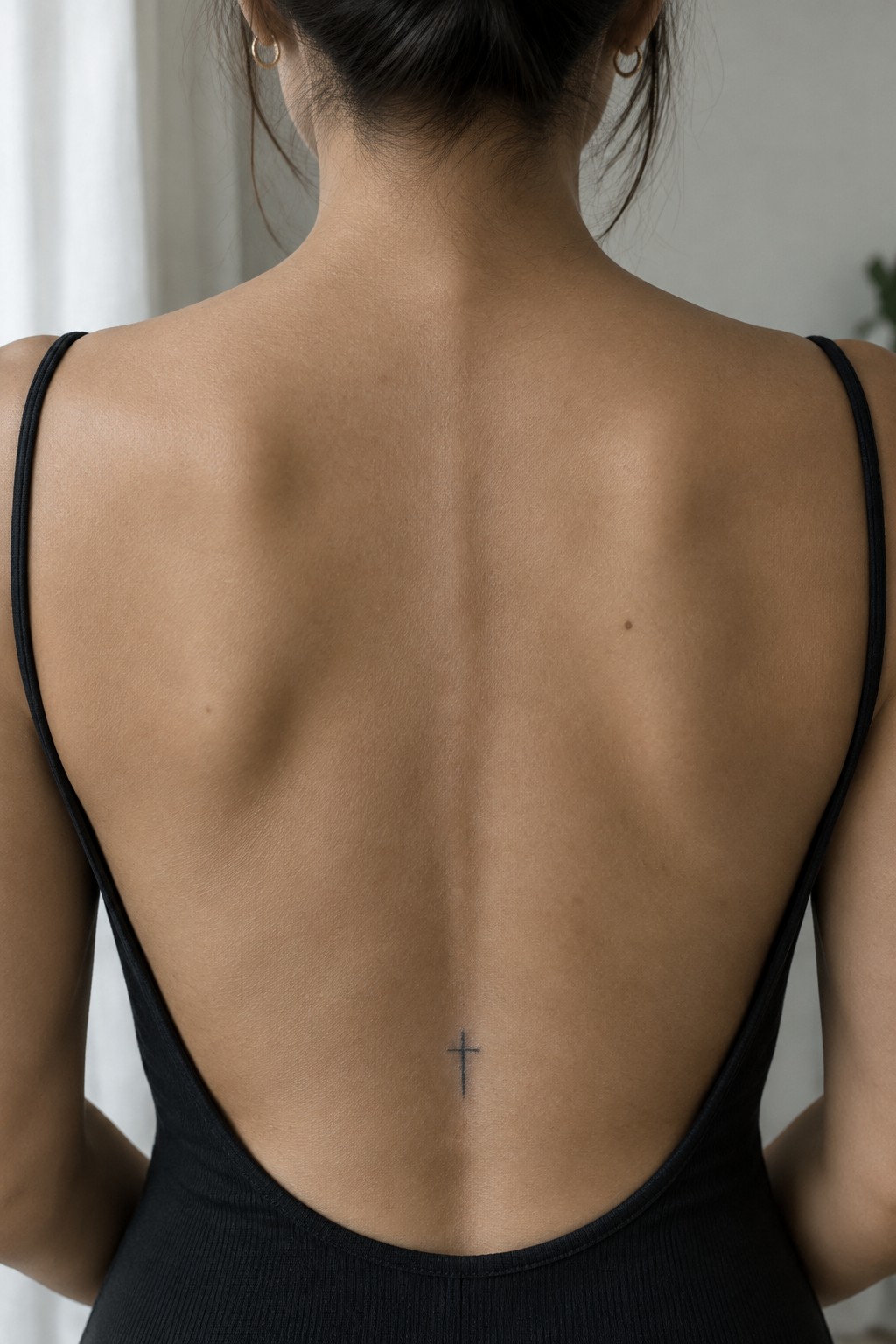 Small fine-line cross tattoo centered on a woman’s lower back above a black open-back top.