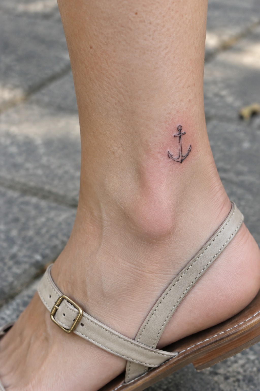 Small black anchor tattoo on the outer ankle above a beige sandal.