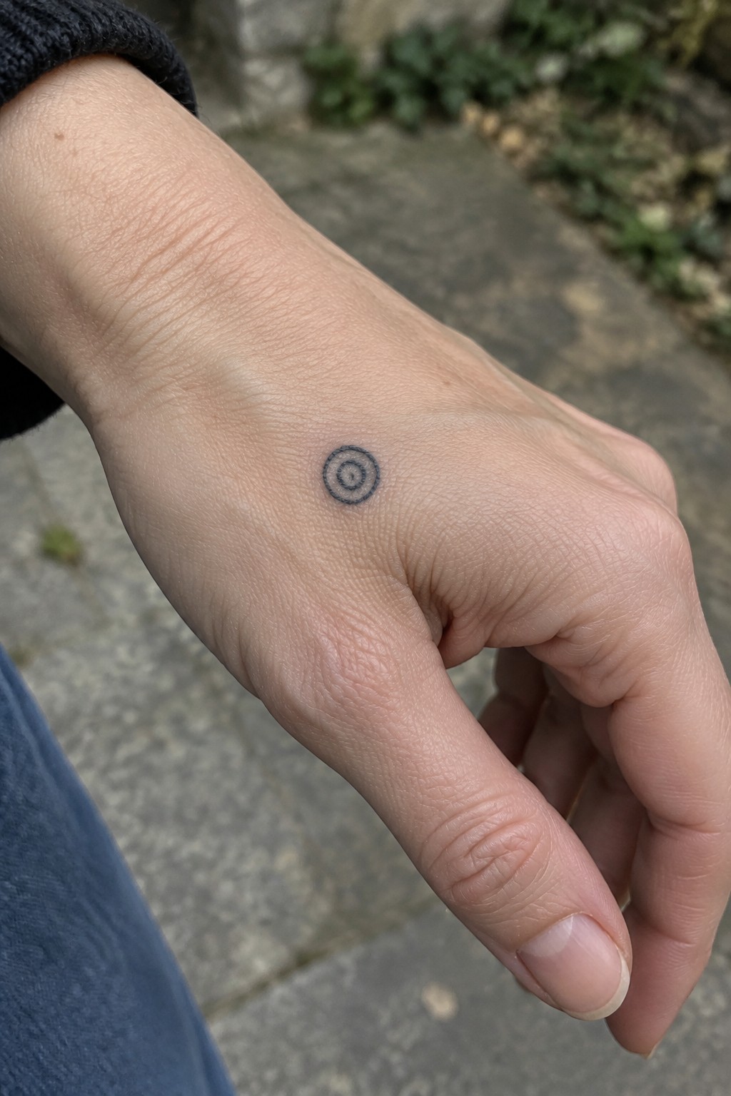 Small black concentric circle tattoo on the back of a hand near the thumb.