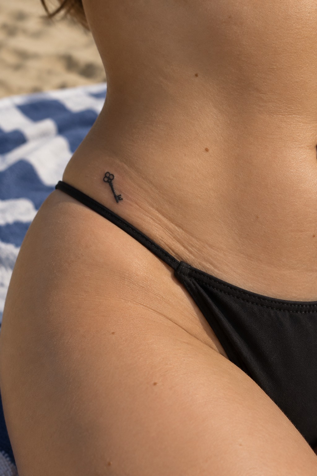 Tiny black key tattoo above the hip near the bikini line, featuring a small floral detail at the top.