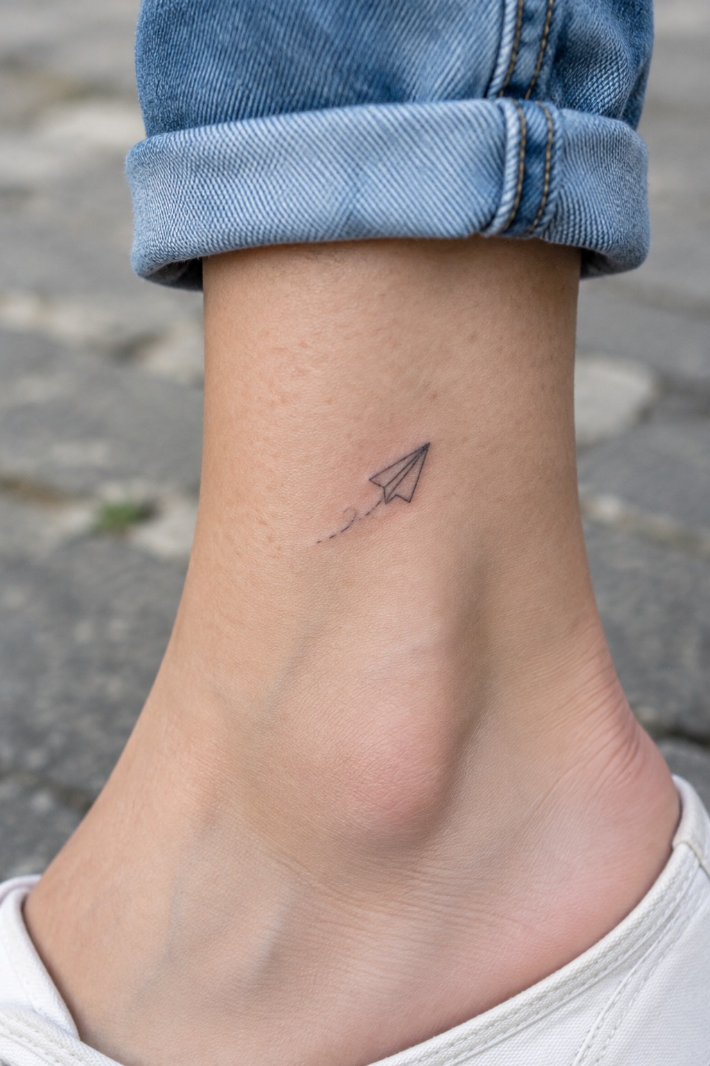 Small black paper airplane tattoo with a dotted trail above the ankle, beneath a rolled-up denim cuff.
