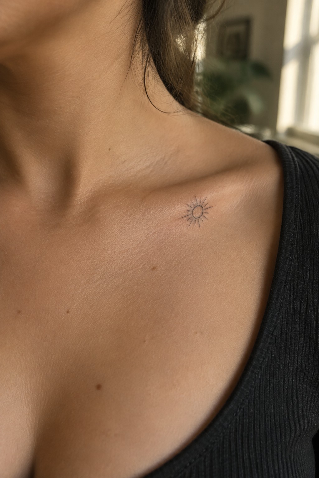 Small fine-line sun tattoo with a circle and thin rays on the upper chest just below the collarbone.
