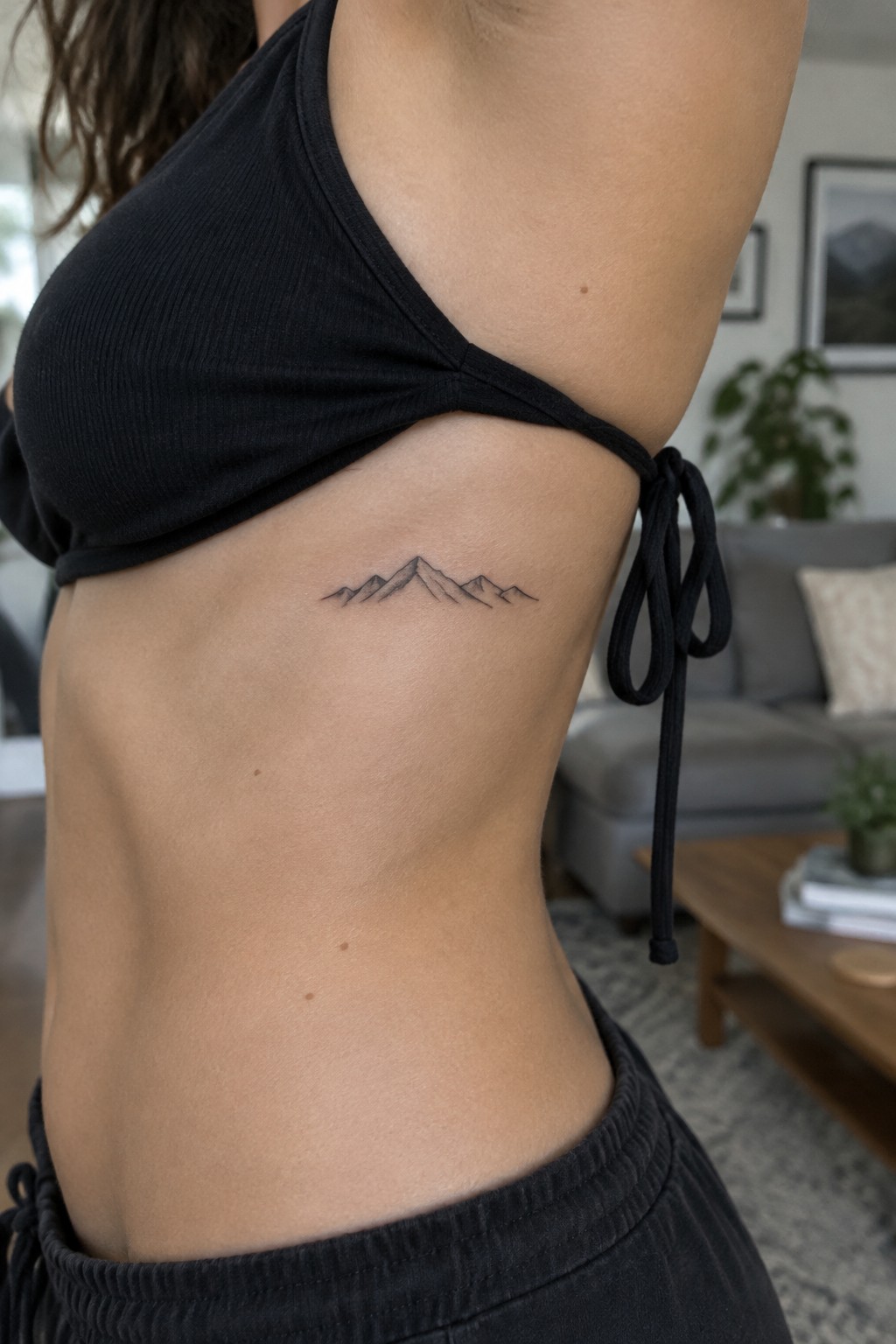 Small minimalist mountain range tattoo in black line art on the ribcage of a woman in a black crop top.