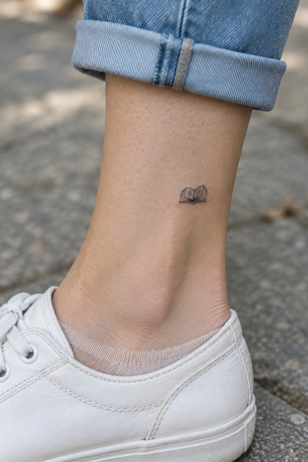 Small open book tattoo on the outer ankle above a white sneaker and light sock