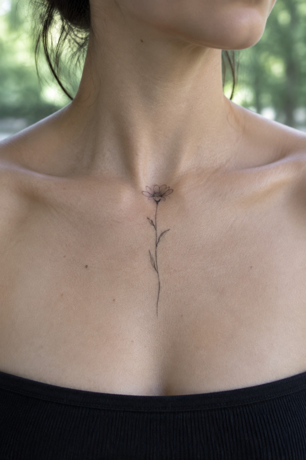Minimalist black line flower tattoo on upper chest above black top with blurred greenery