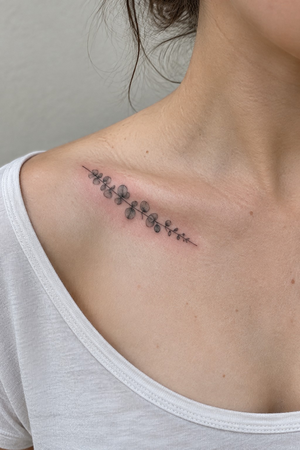 Minimalist fine line tattoo of a thin horizontal stem with small rounded leaves on the collarbone area