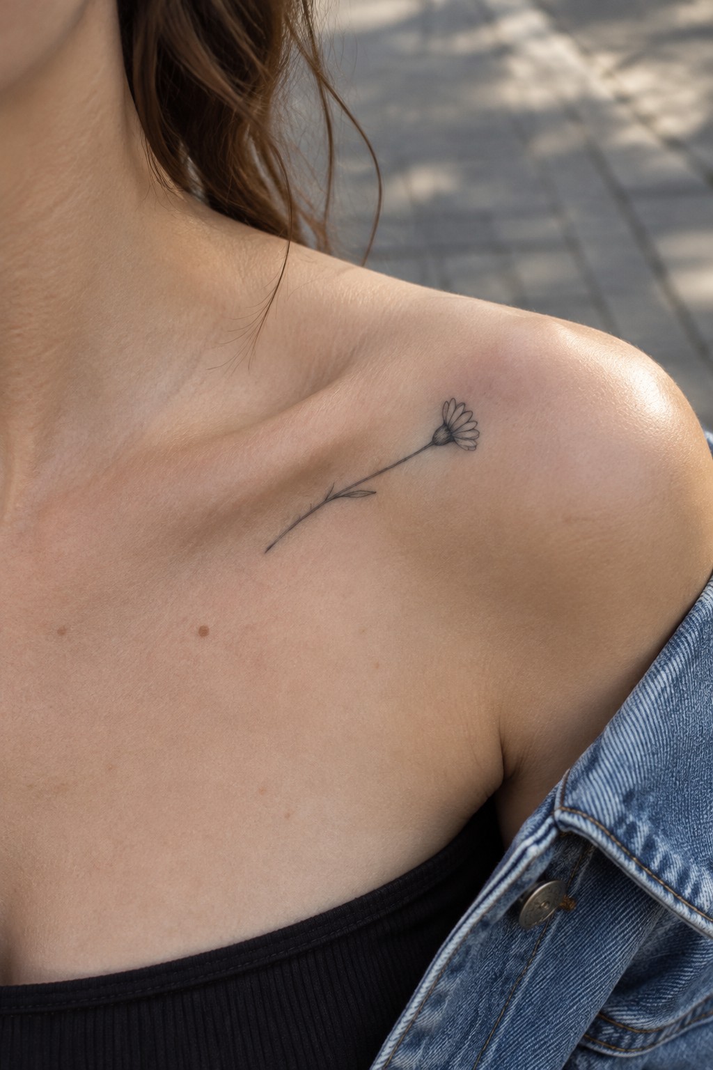 Small daisy tattoo on woman's shoulder over black top and blue denim jacket.