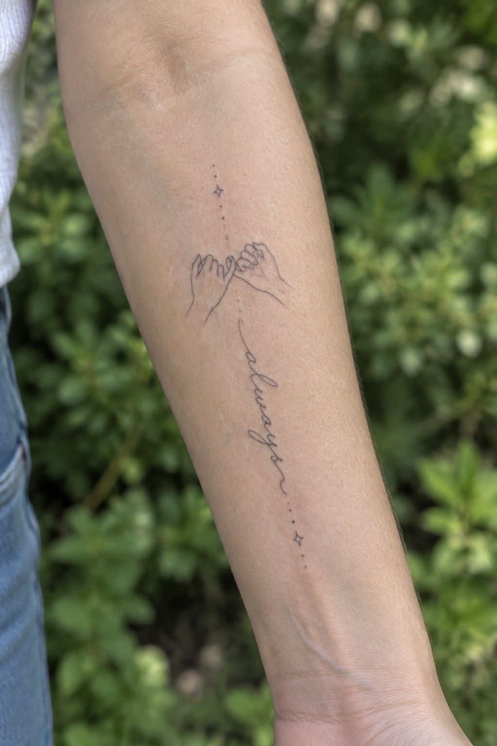 Forearm tattoo of linked pinky hands, stars, and cursive 