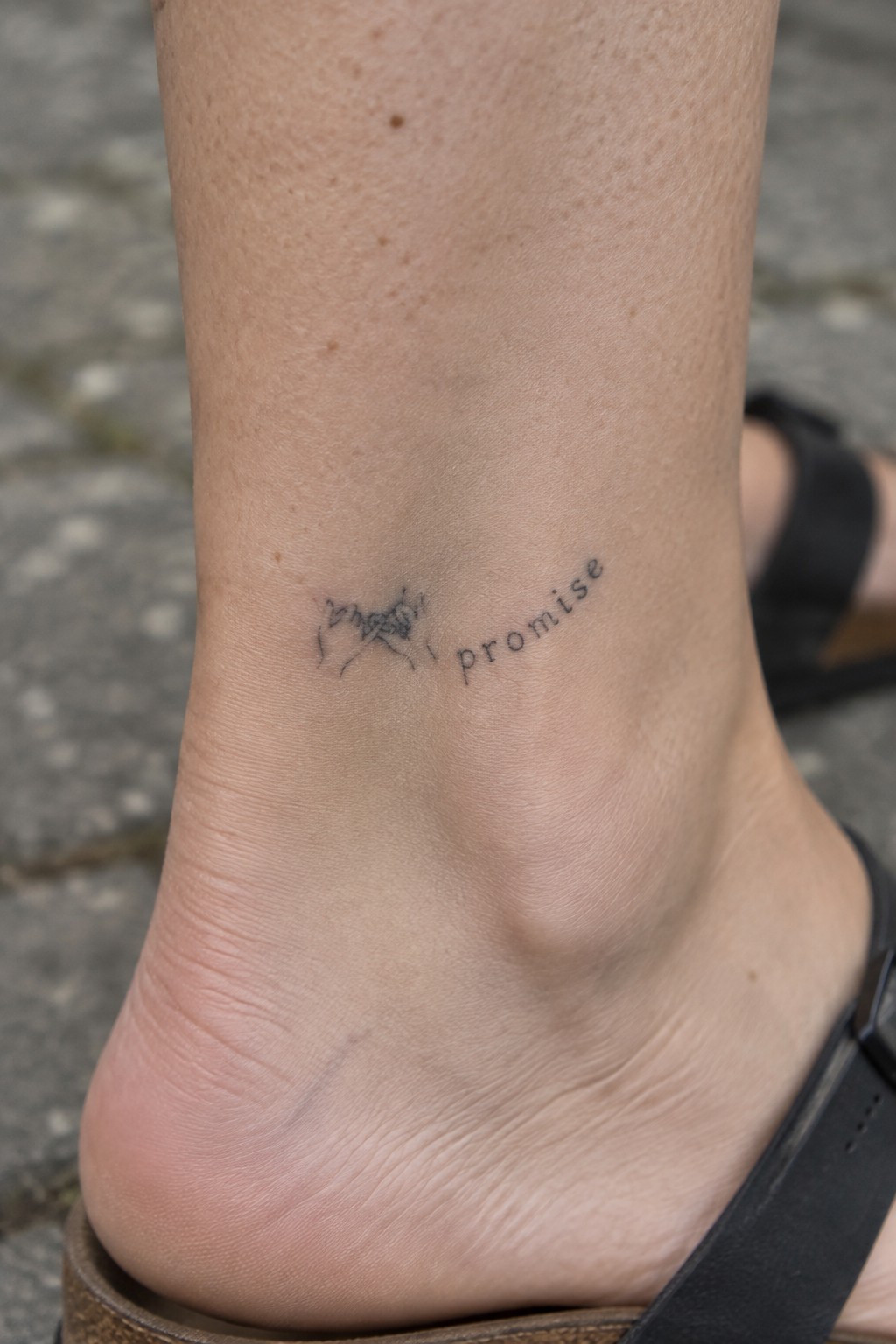 Fine line black tattoo of two linked pinky fingers beside the word promise on an ankle