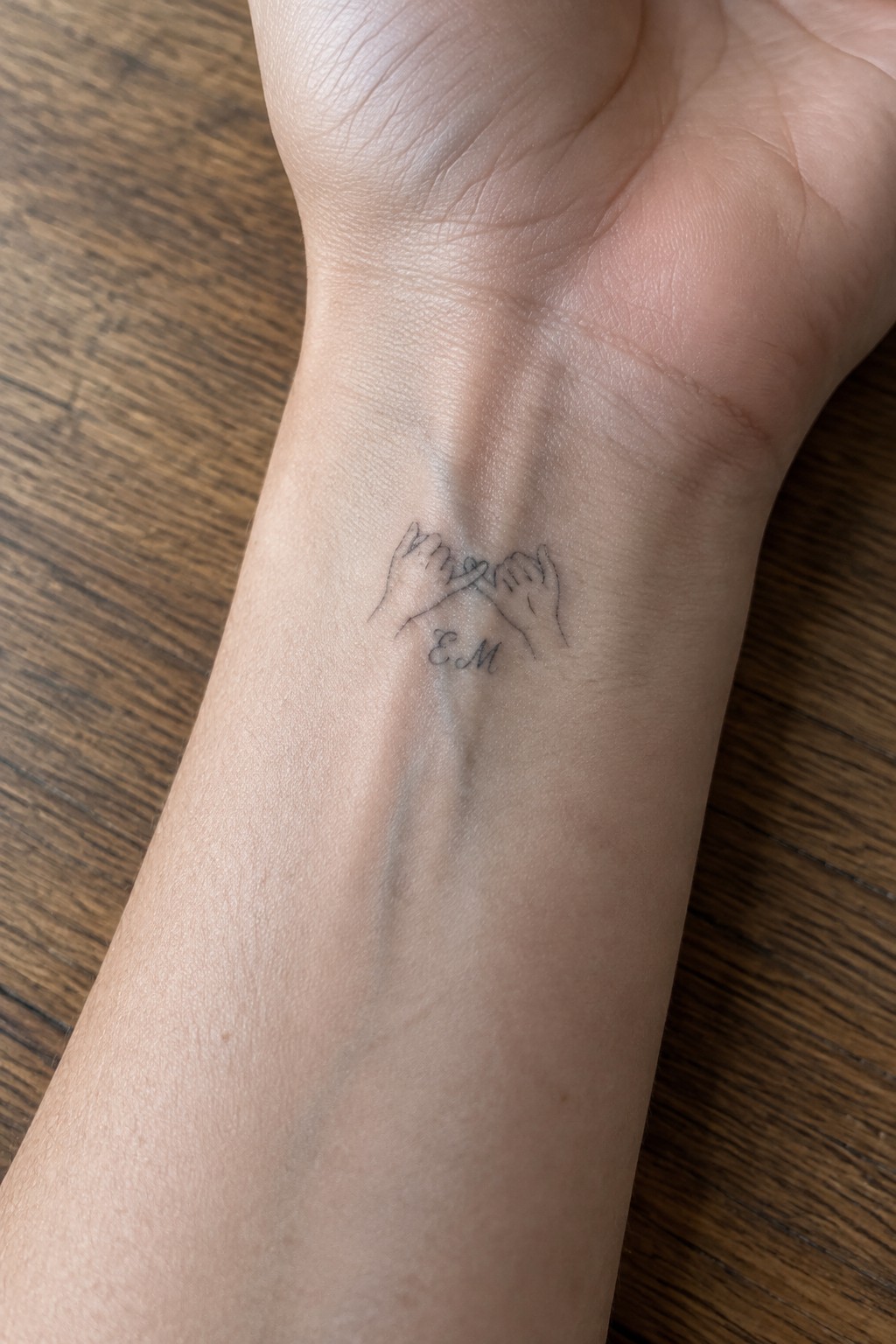 Fine line tattoo of two linked pinky fingers with EM initials on the inner wrist