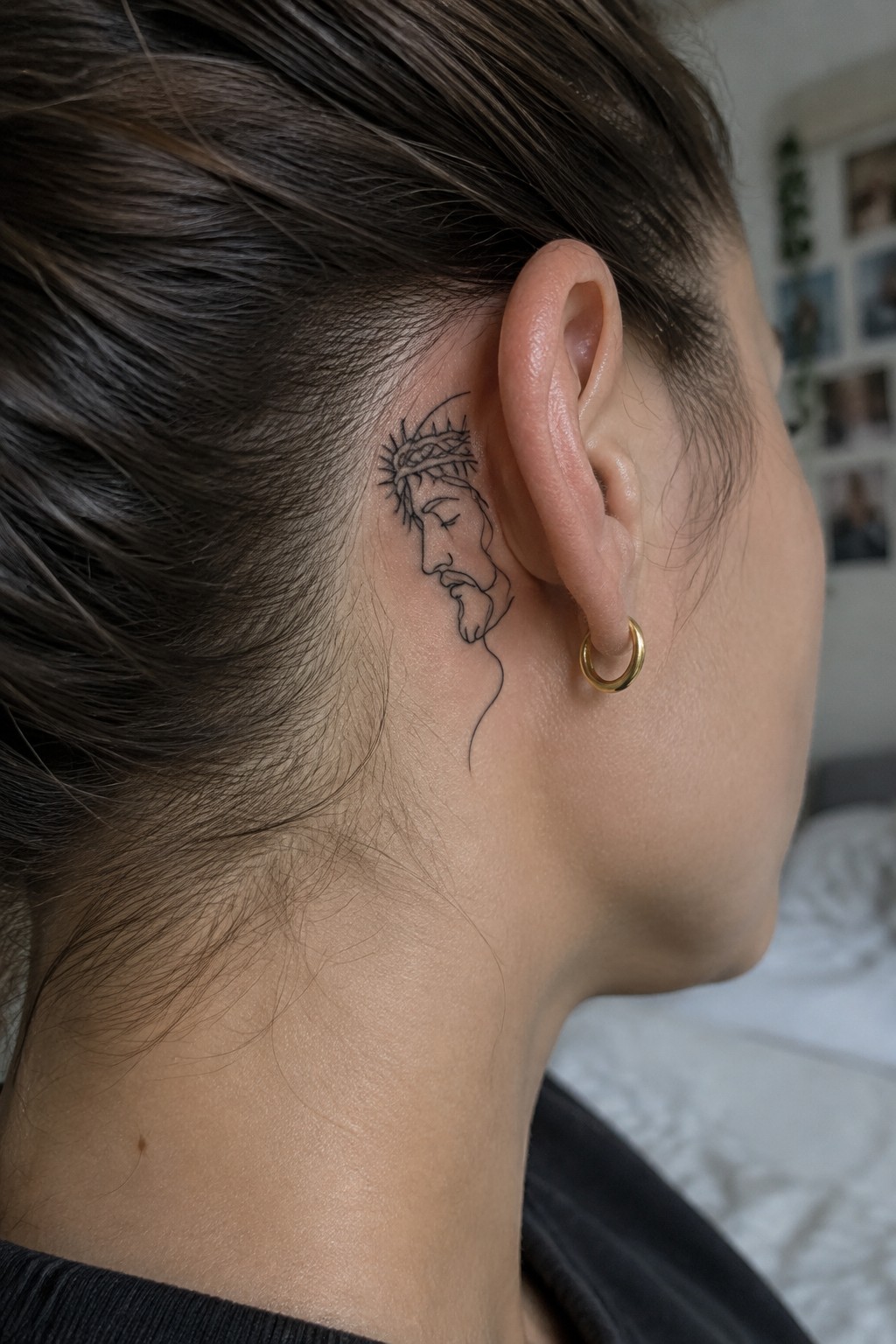 Minimalist line art tattoo of Jesus in profile wearing a crown of thorns, placed behind the ear on the side of the head.