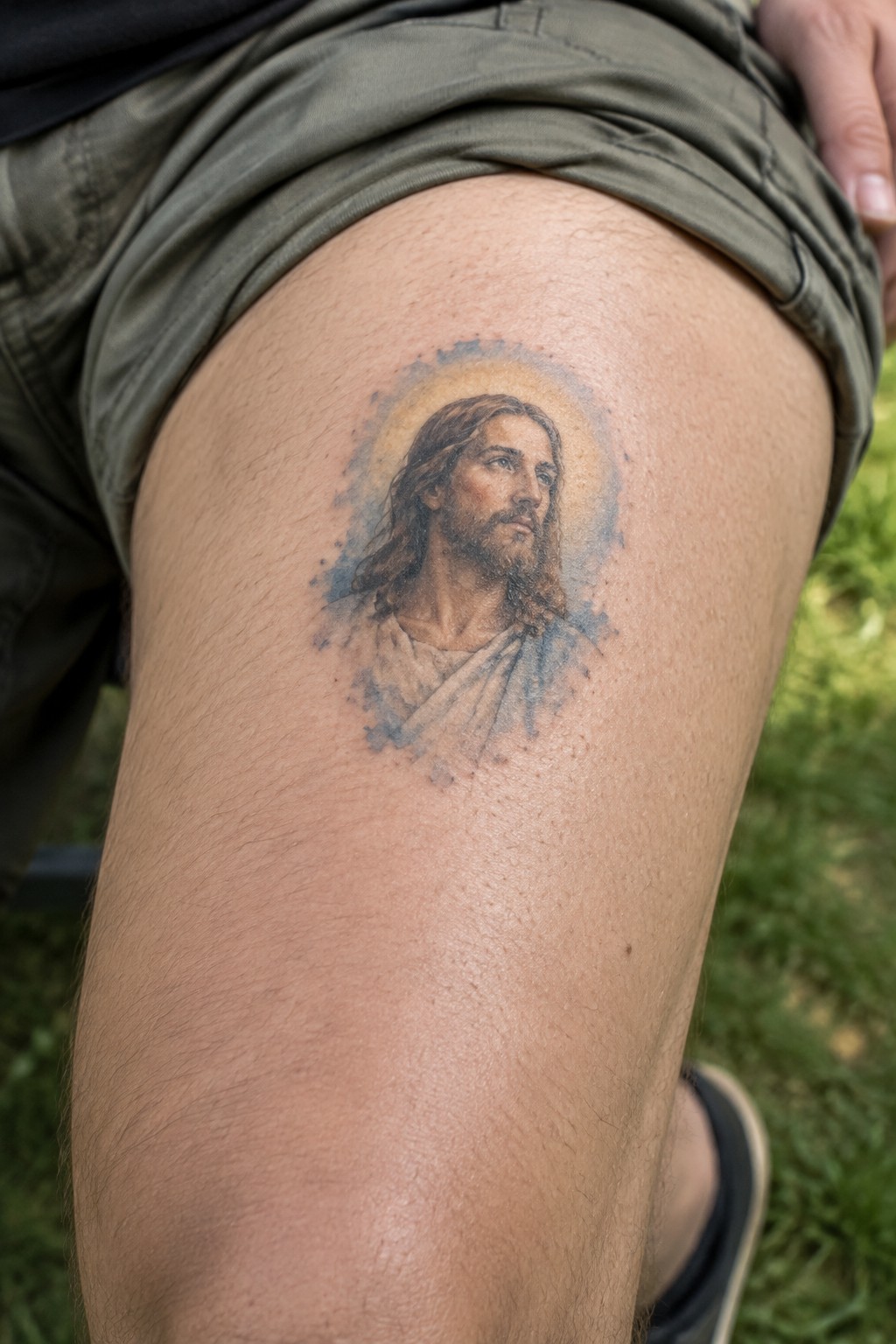 Jesus tattoo with halo on thigh, blue watercolor splashes, green shorts, grass background.