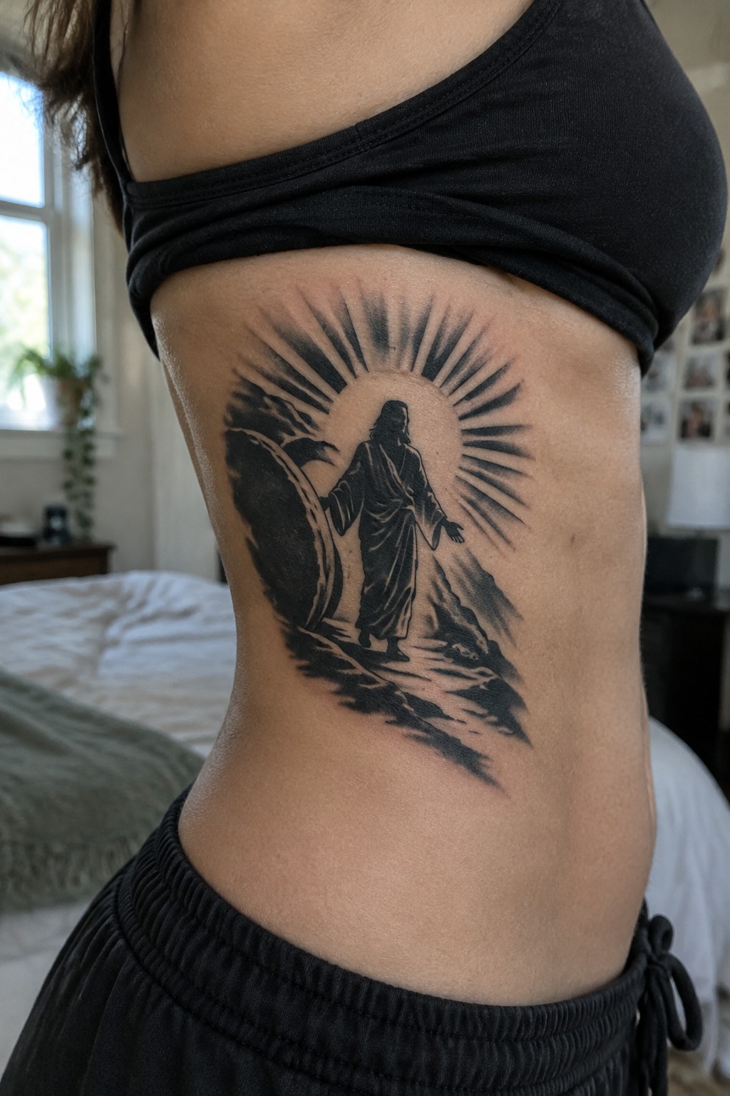 Black ink tattoo of robed figure with sun rays on bare torso in black top