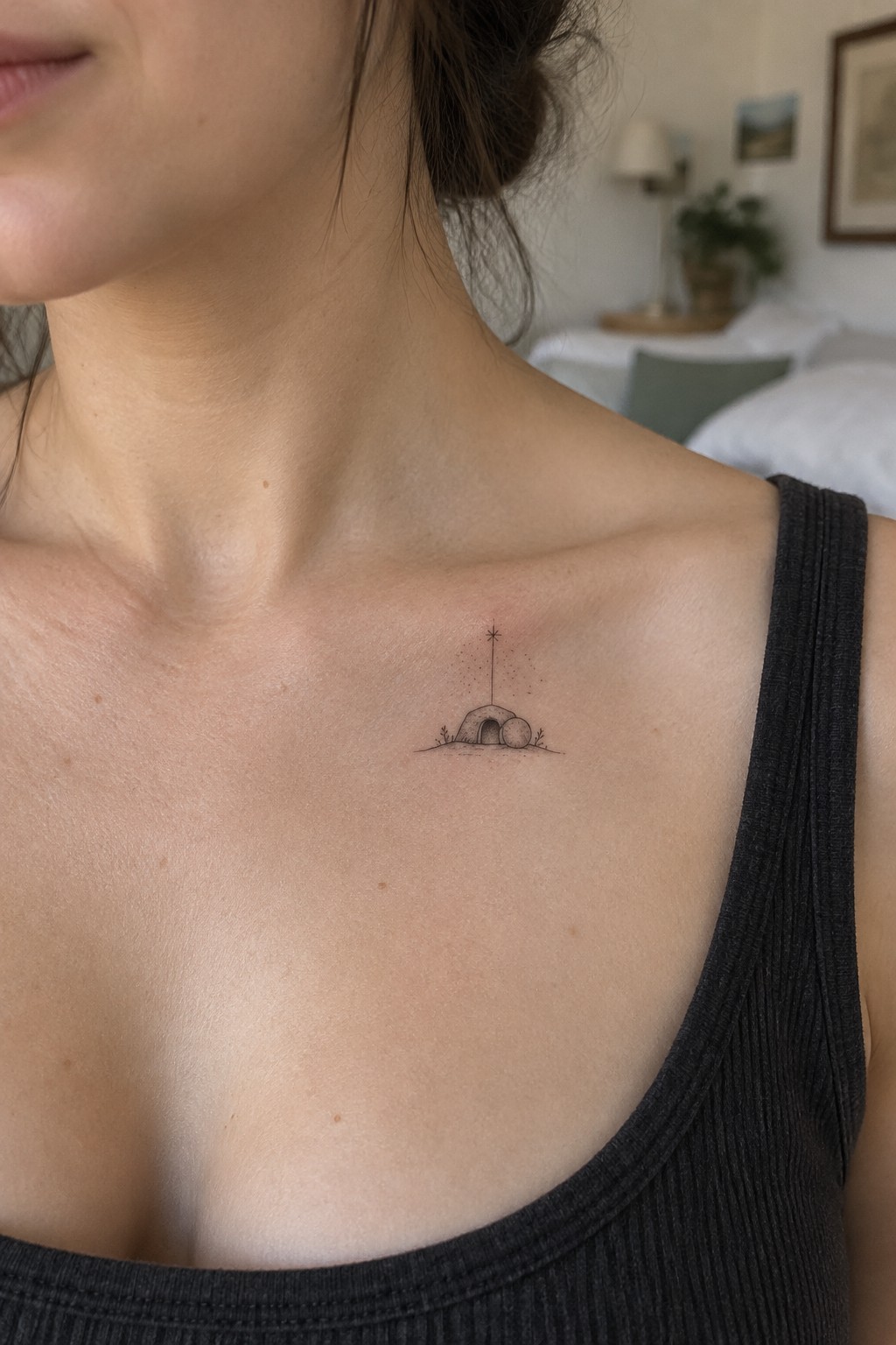 Small black tattoo of a domed hut under a star on woman's upper chest in gray tank top