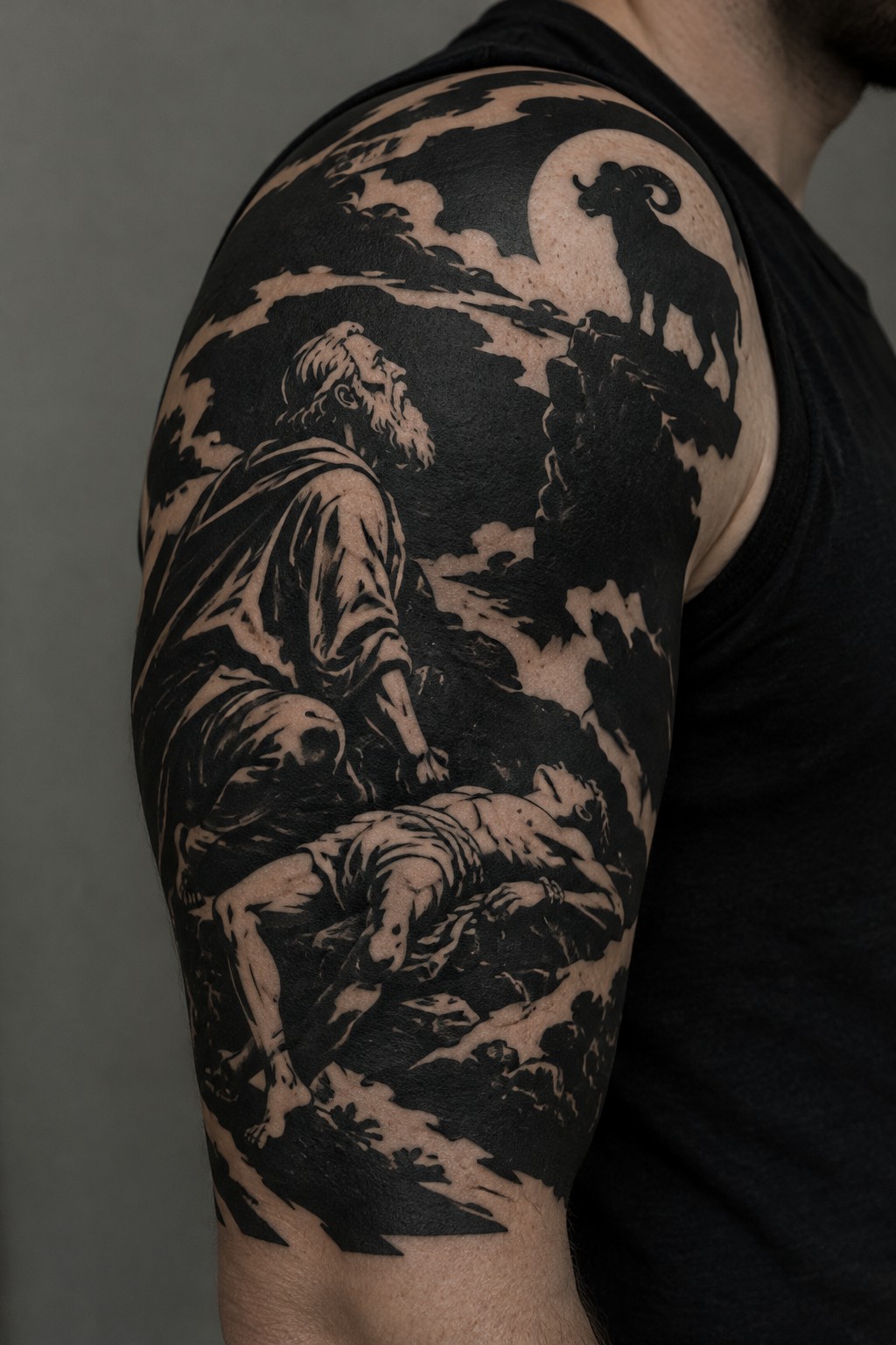 Black ink tattoo on shoulder shows robed man, bound figure, ram on cliff under full moon