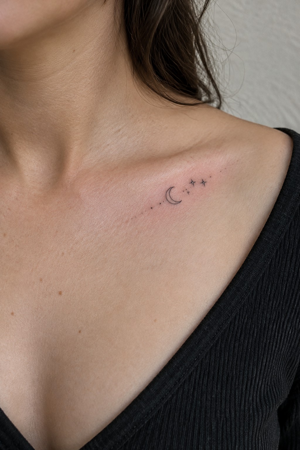 Small fine-line crescent moon, stars, and dots tattooed along a woman’s collarbone.