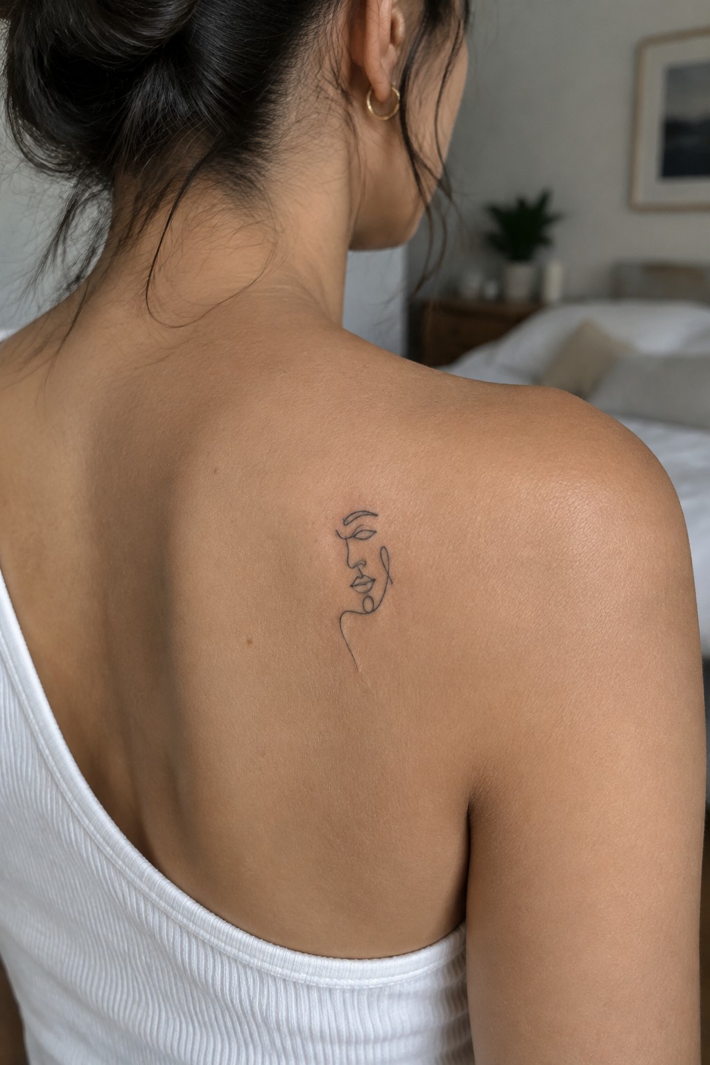 Small fine-line tattoo of a woman’s face outline on the upper back of a person’s shoulder.