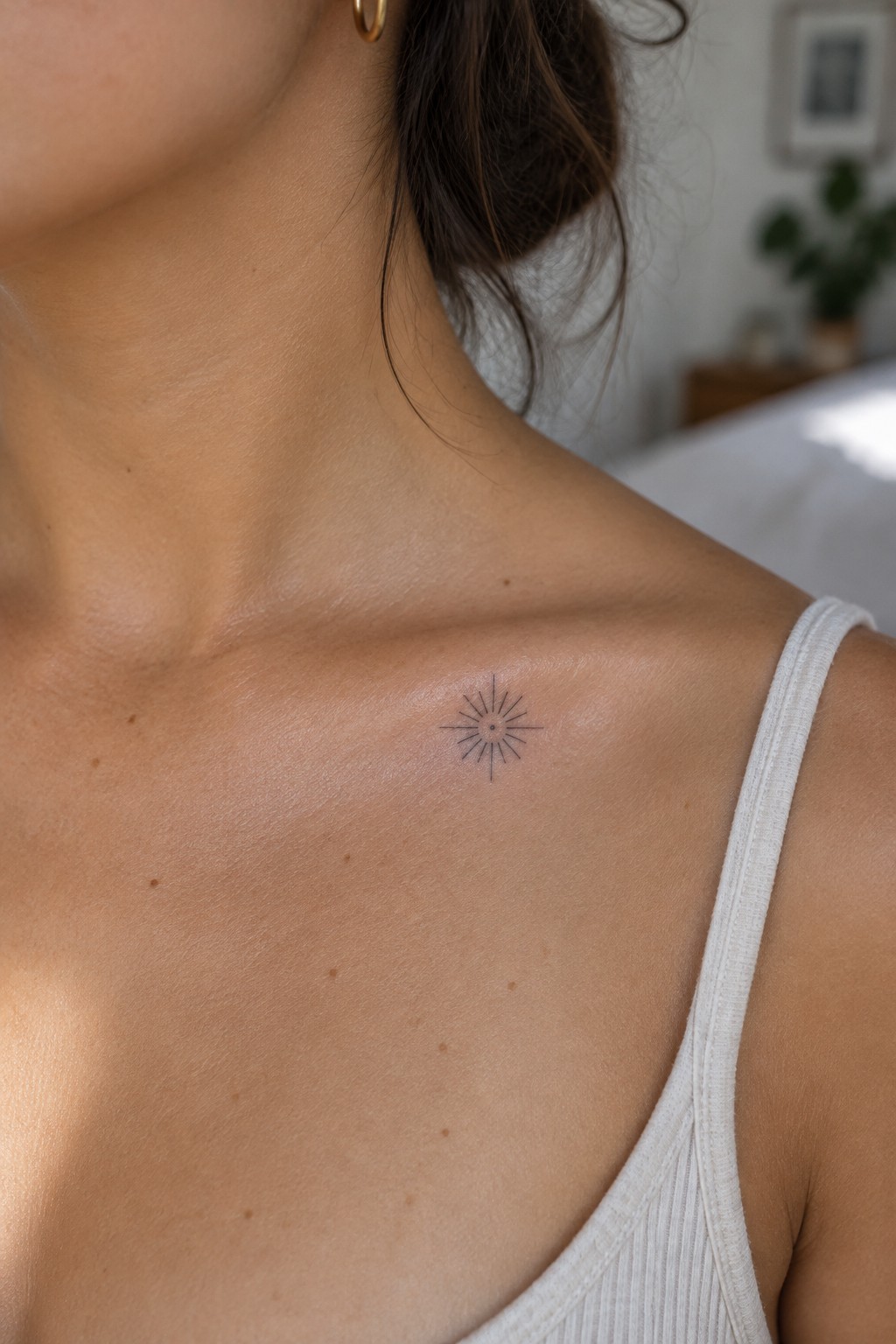 Small fine-line sunburst tattoo on a woman’s collarbone, just above a white tank top.