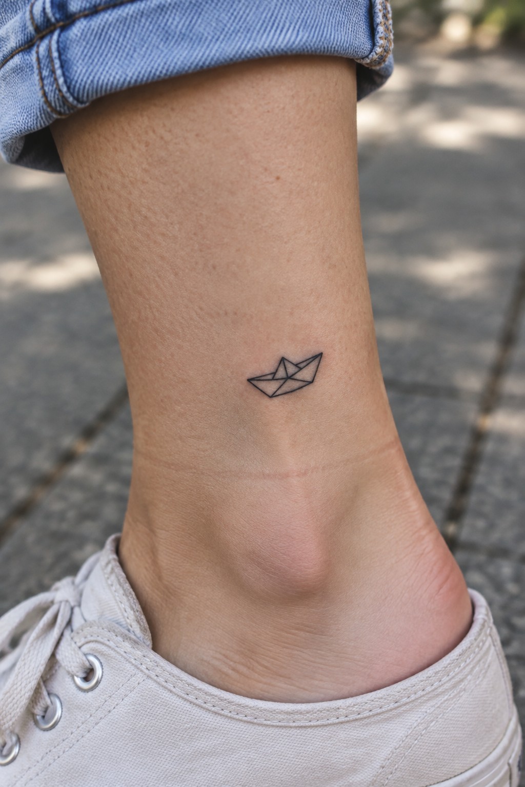Small black linework paper boat tattoo on the outer ankle above a white sneaker.