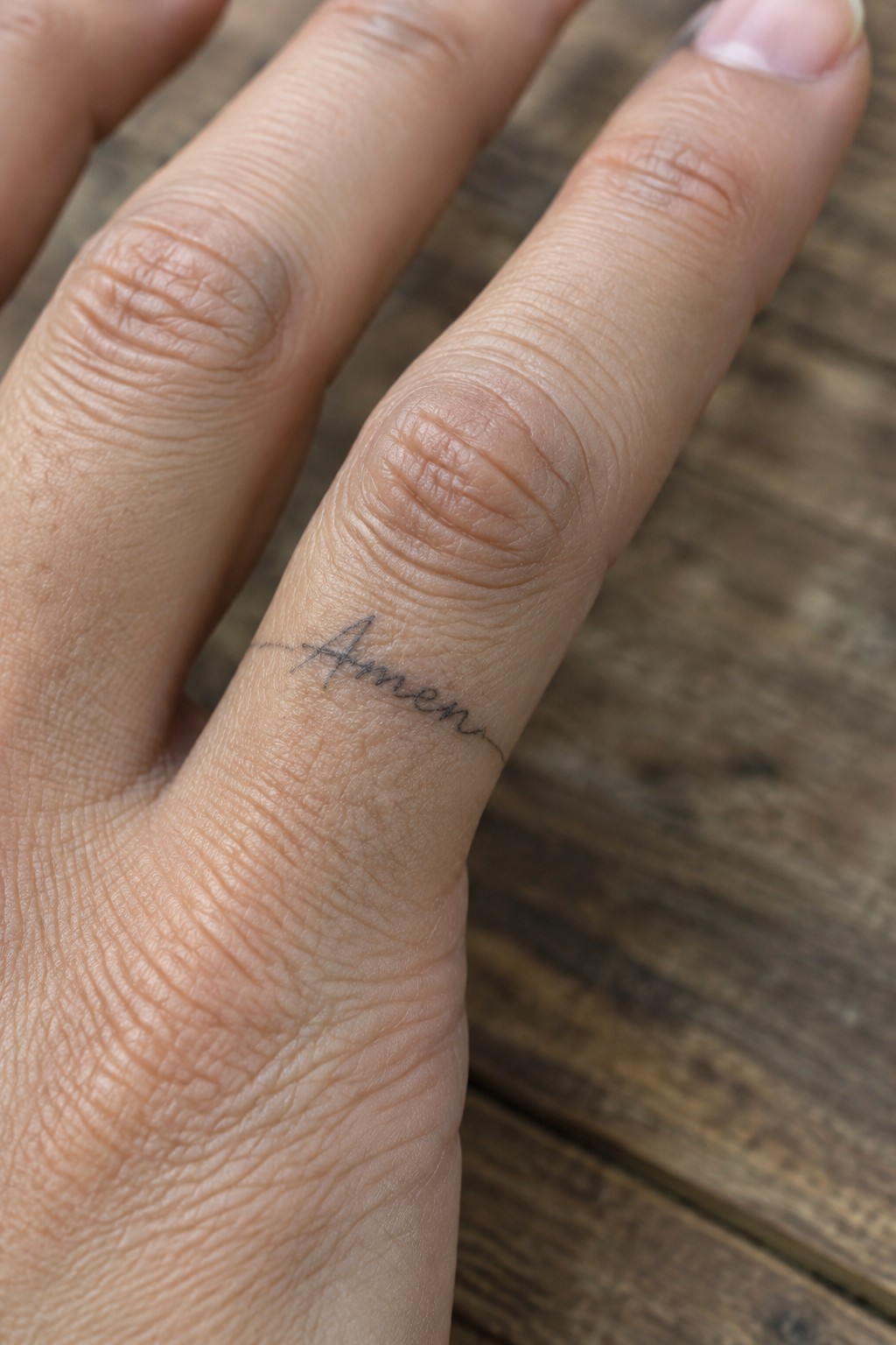 Close-up of a fine line amen tattoo in cursive script on the side of a finger resting on wood.