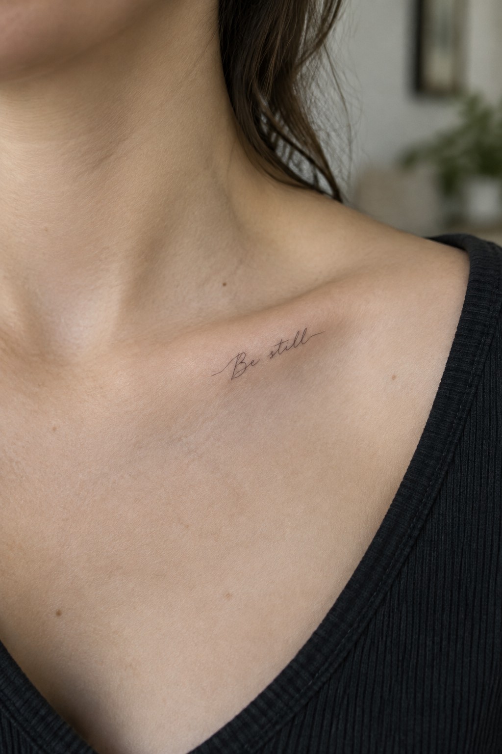 Script 'Be still' tattoo on collarbone above black ribbed top on light skin