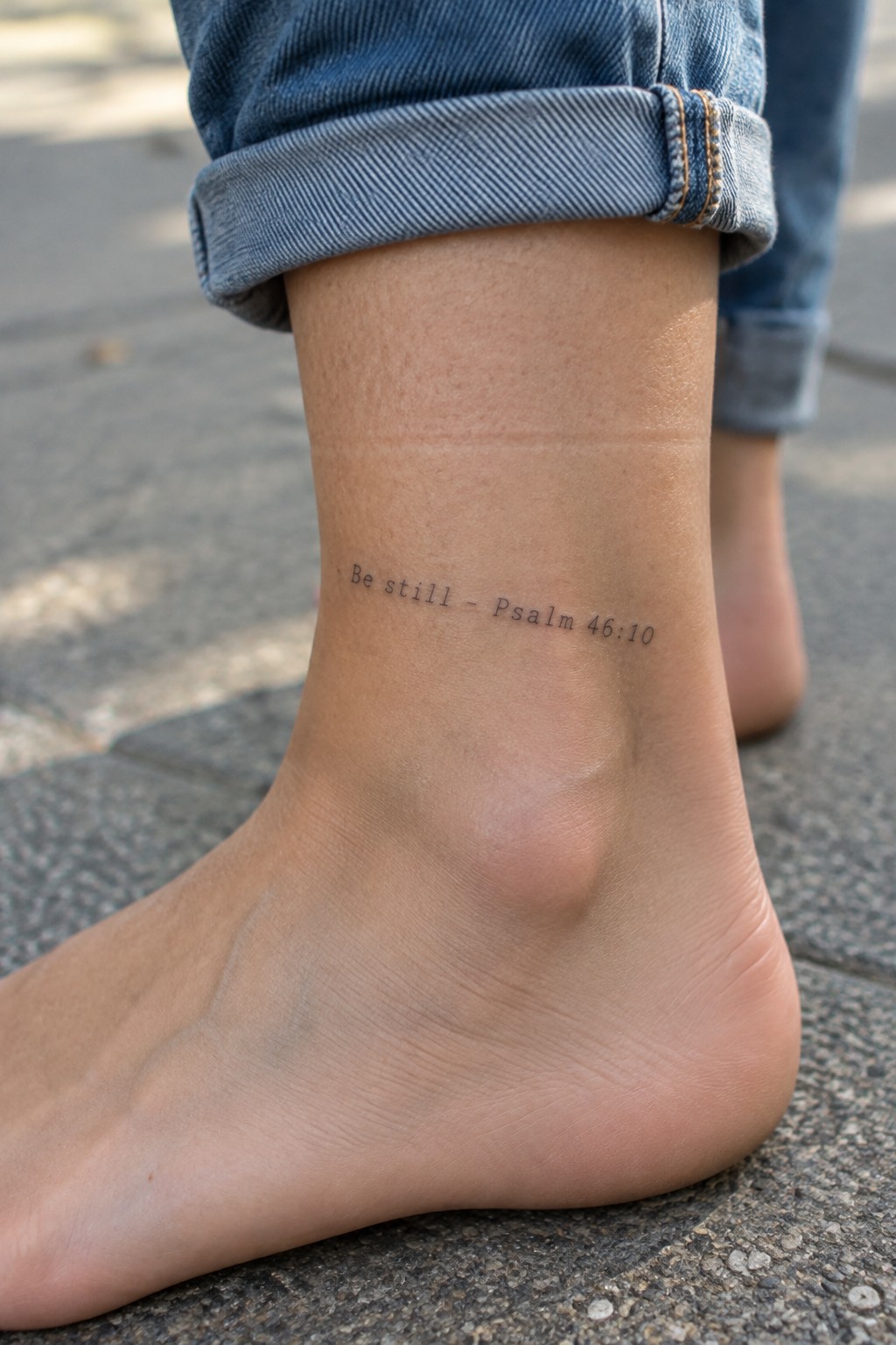 Bare foot with 'Be still - Psalm 46:10' tattoo on ankle under cuffed blue jeans.