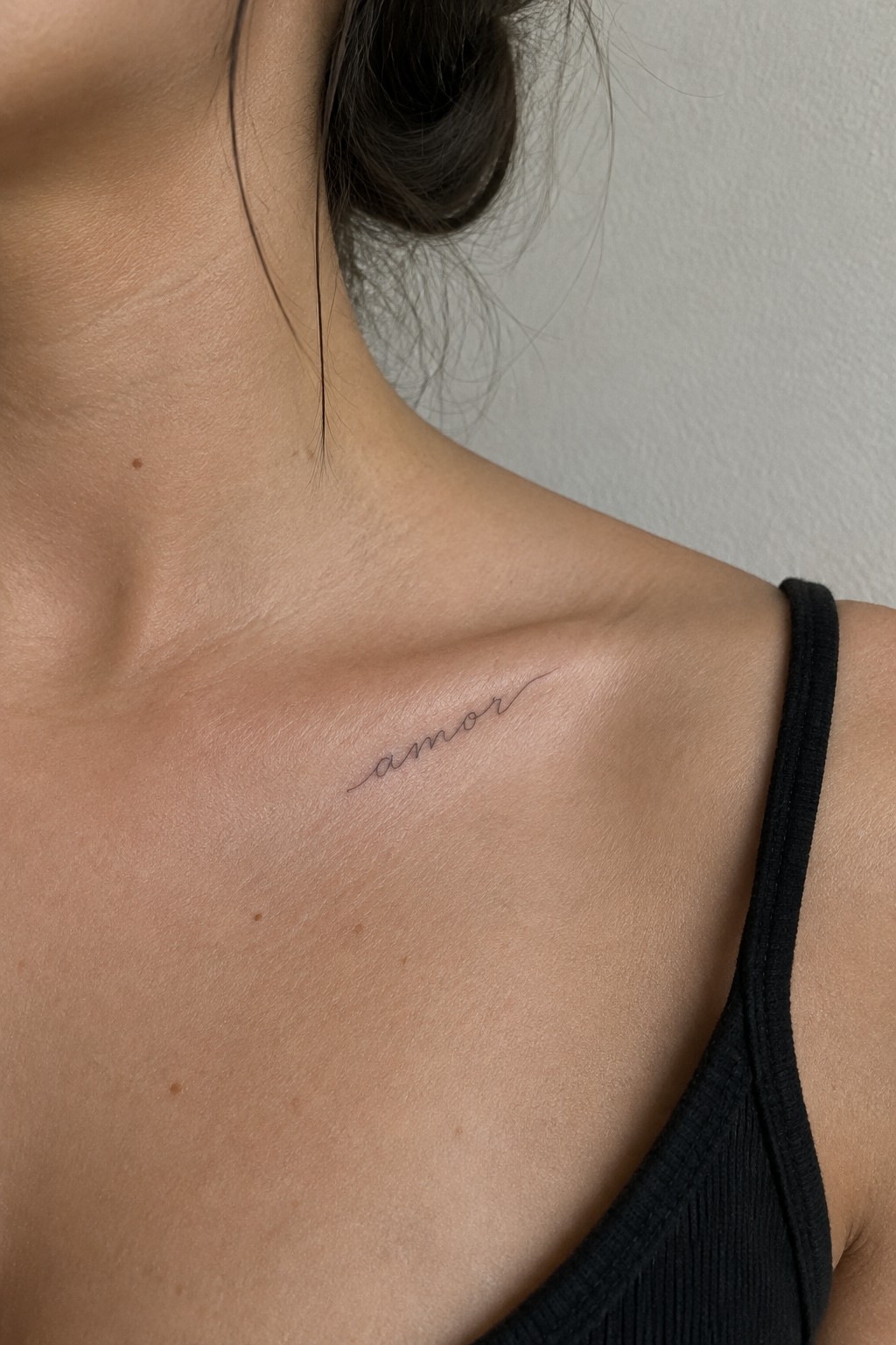 Small fine-line “amor” script tattoo angled below a woman’s collarbone.