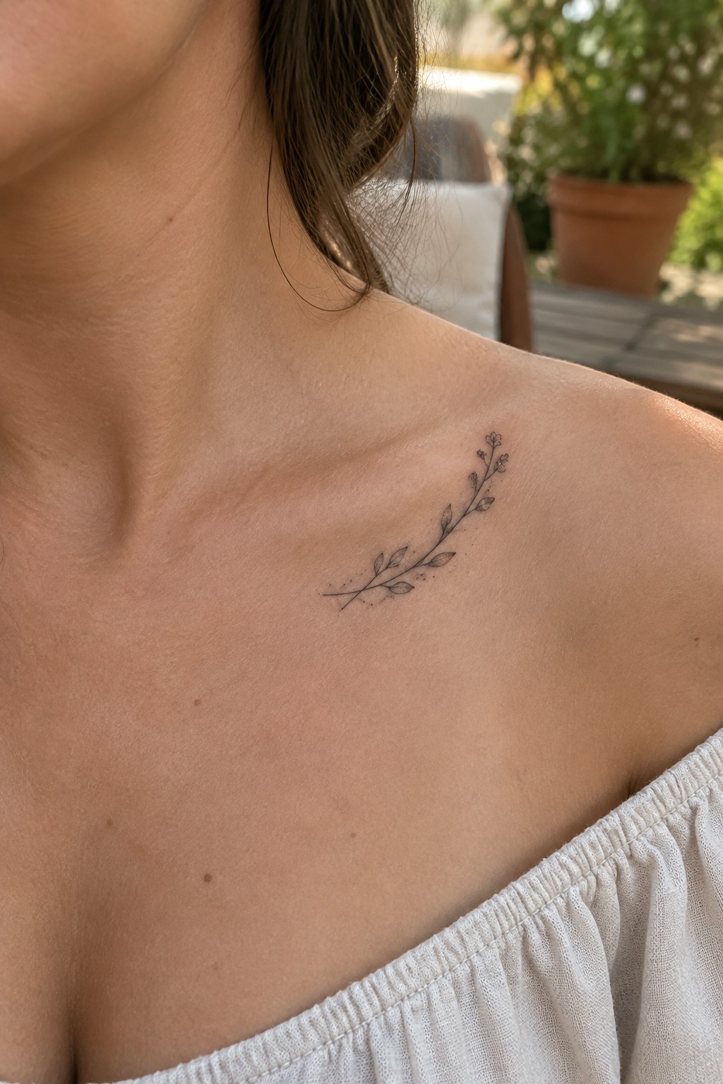 Fine-line tattoo of a curved leafy branch with small buds on a woman’s upper chest near the collarbone.