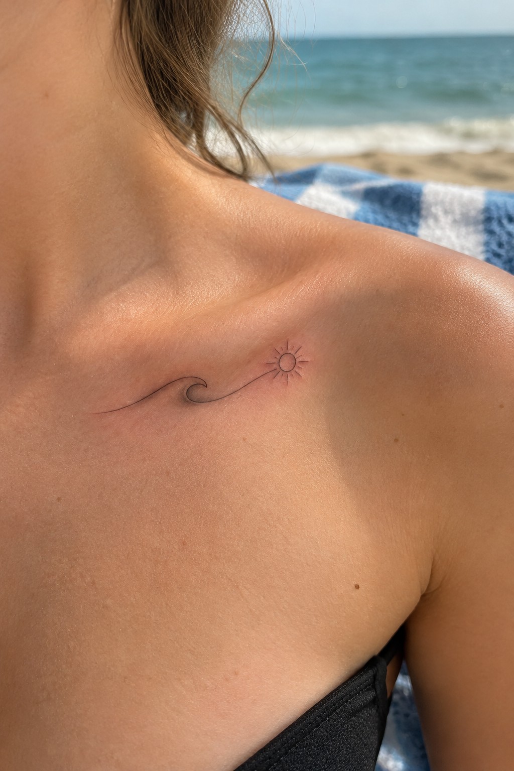 Fine-line tattoo of a small wave flowing into a sun on a woman’s upper chest below the collarbone.