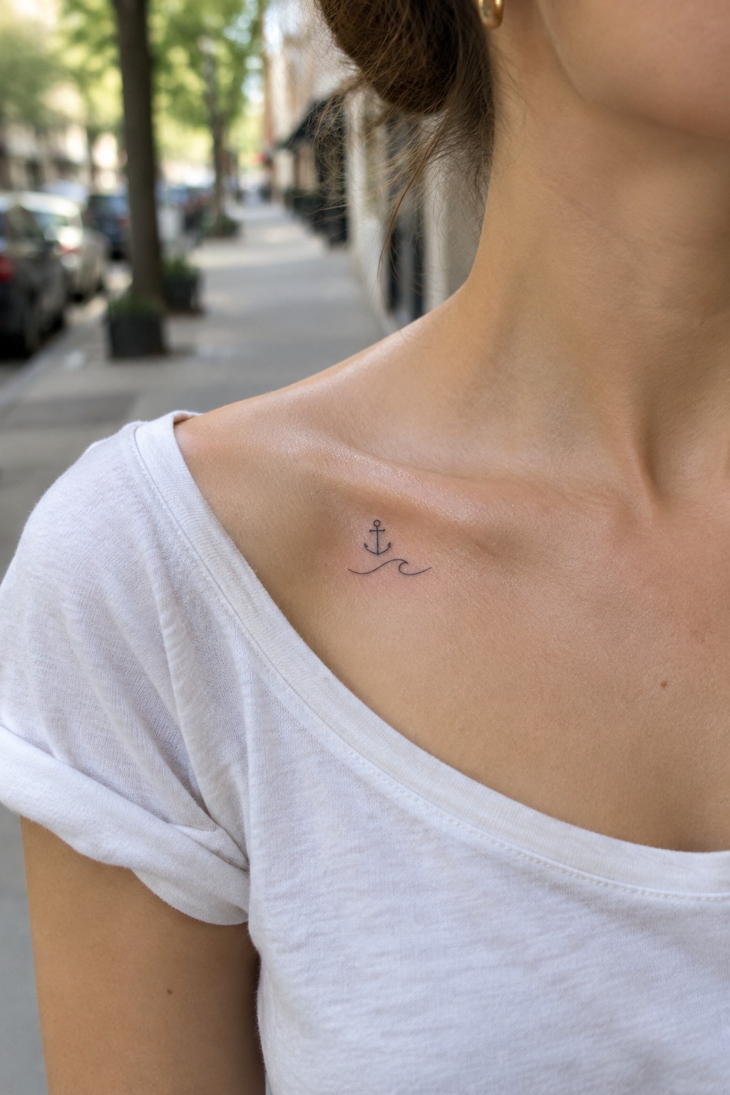 Small fine-line anchor and wave tattoo below a woman’s collarbone.