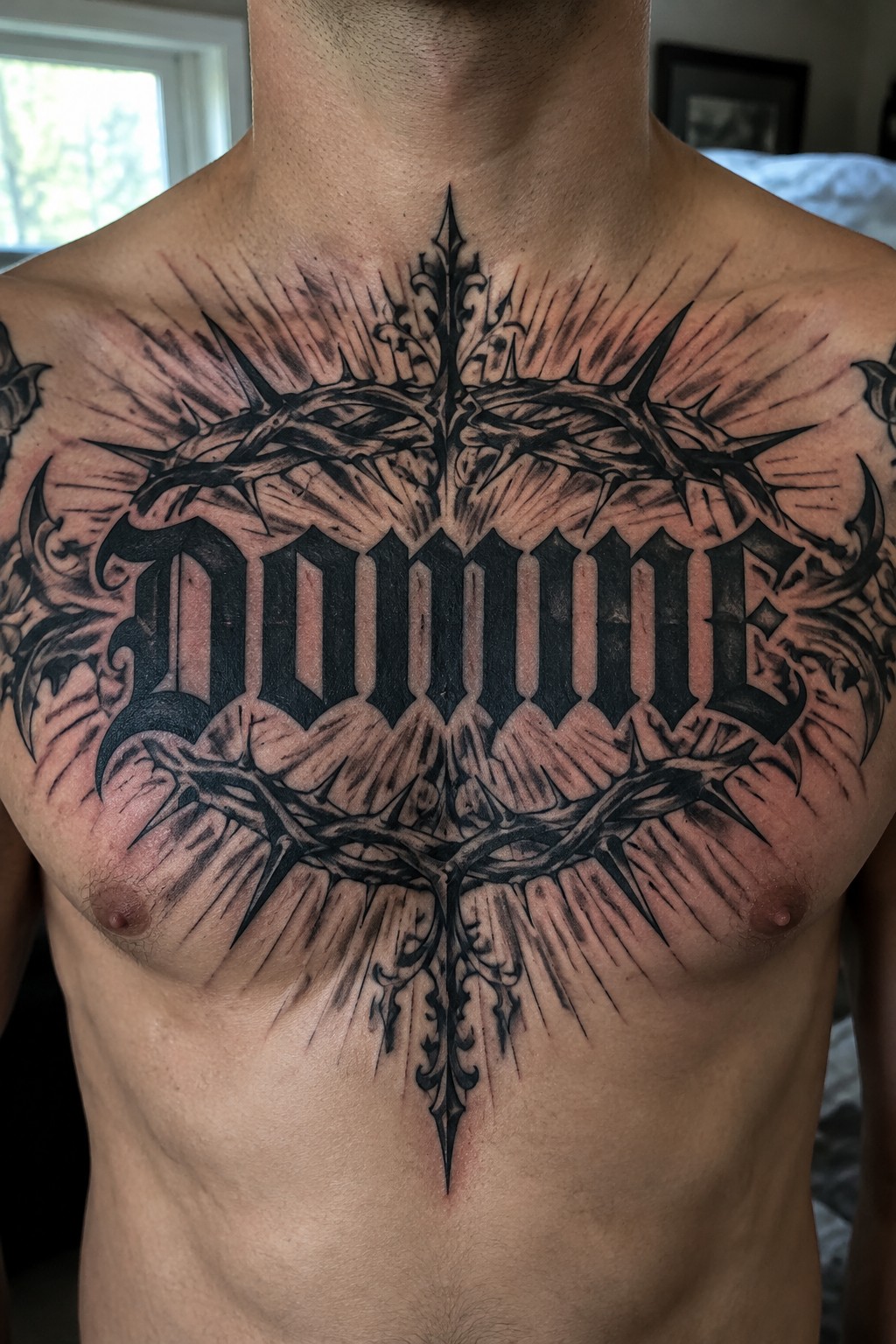 Large black gothic tattoo on a man's chest reading Domine, surrounded by crown of thorns and radiating lines in dark shading.