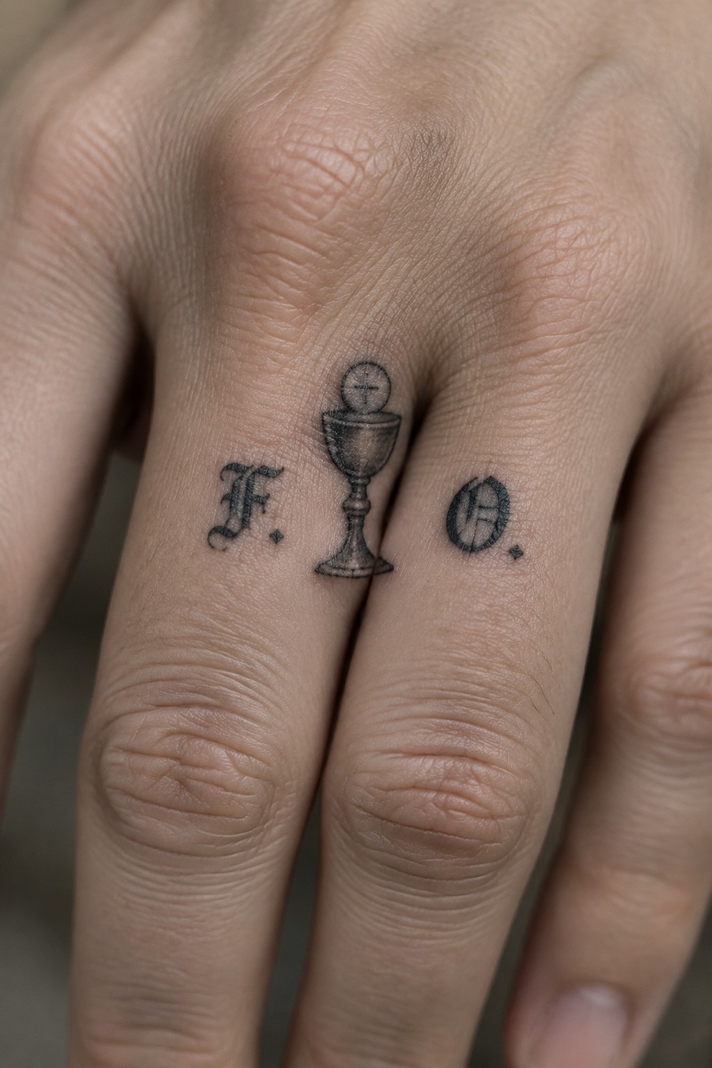 Fingers with black tattoos of F., cross-topped chalice, and O. on skin.