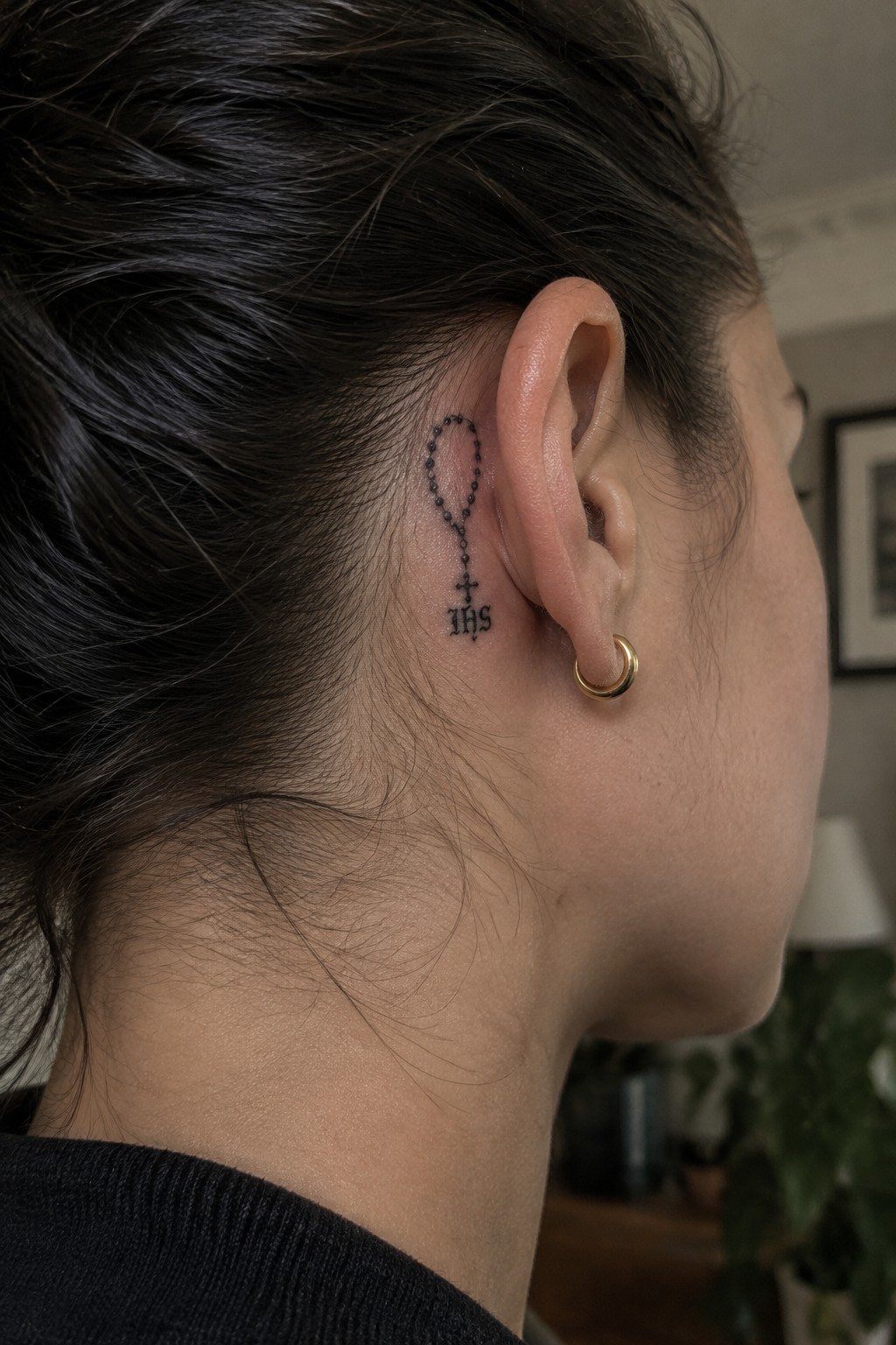 Small black rosary tattoo with beads, cross, and IHS lettering placed behind the ear