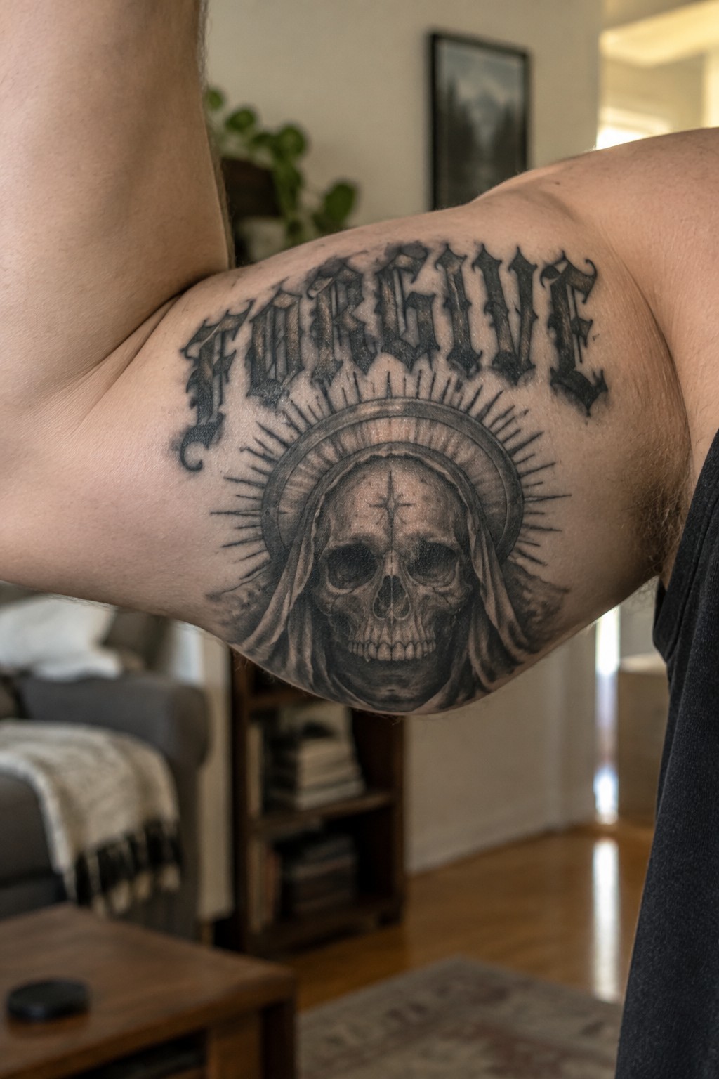 Black and gray tattoo on upper arm with the word FORGIVE in gothic lettering above a hooded skull wearing a veil and a cross on its forehead, surrounded by a radiant halo.