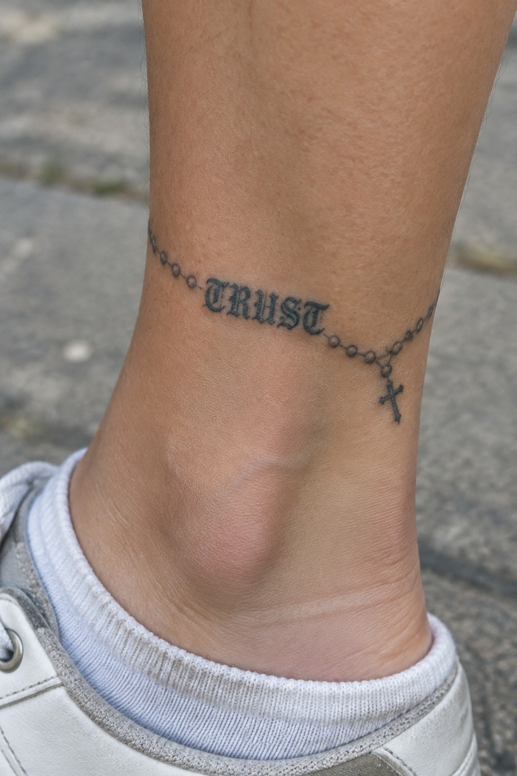 Black rosary tattoo spelling TRUST with cross on ankle over white sock and shoe