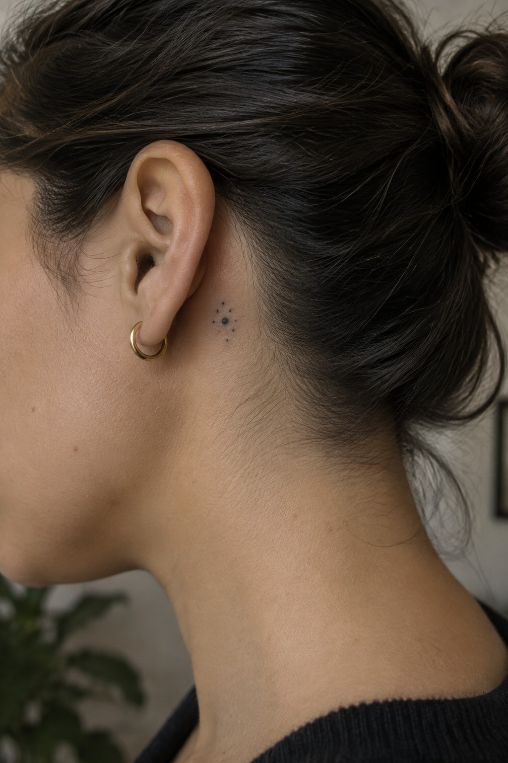 Close-up of a small cluster of black dots tattooed behind the ear on a person with dark hair in a bun.