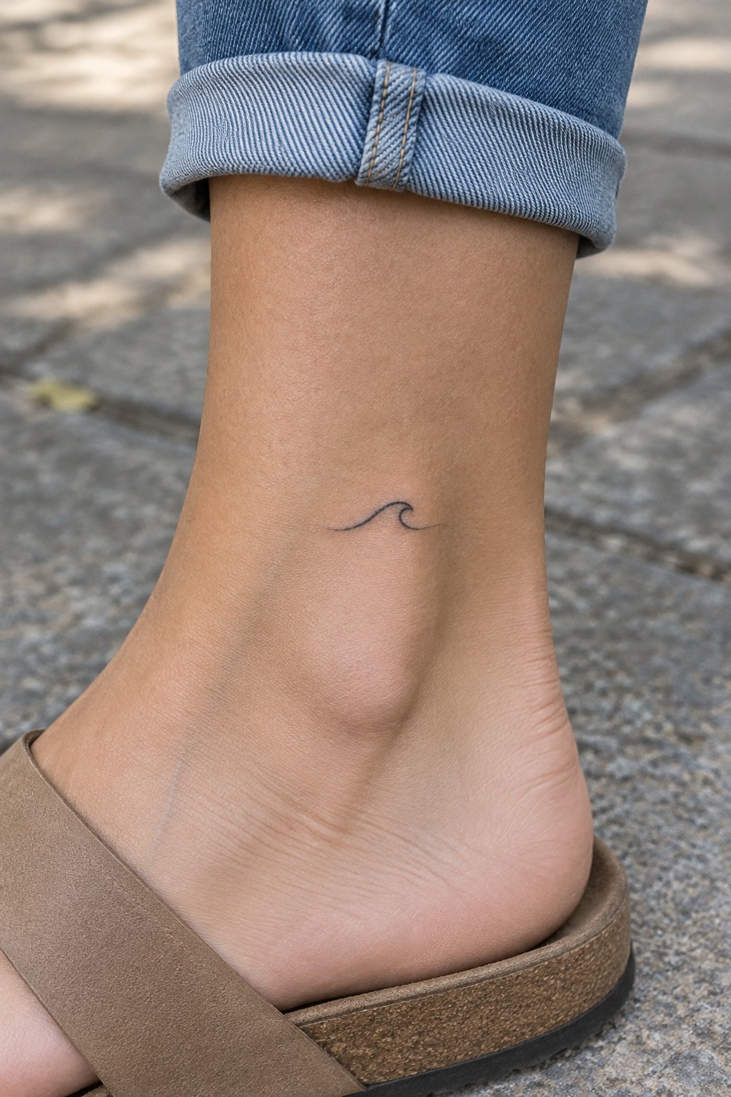 Ankle with small wave tattoo, cuffed blue jeans, and brown sandal on pavement