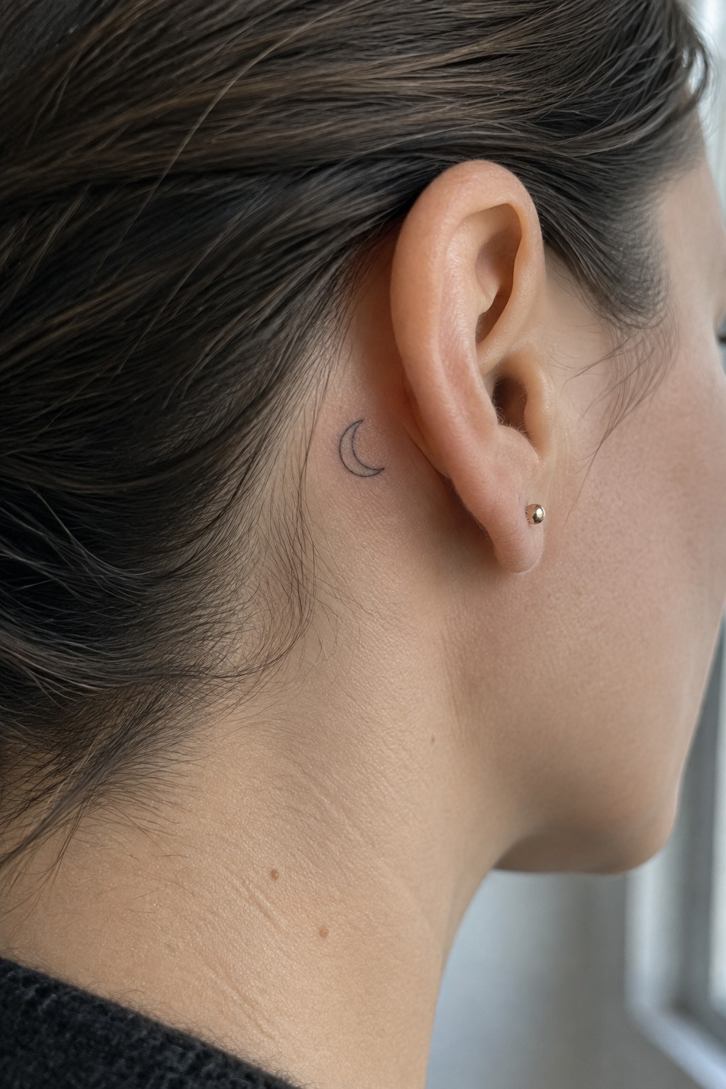 Close-up of ear with crescent moon tattoo and gold stud earring in brown hair