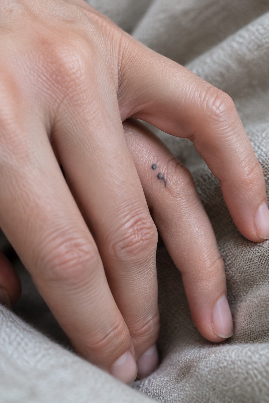 Two hands resting together with a small semicolon tattoo on one finger atop beige fabric.