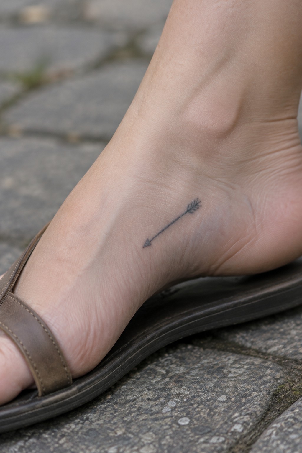 Ankle with small black arrow tattoo above brown sandal strap on gray stone pavement