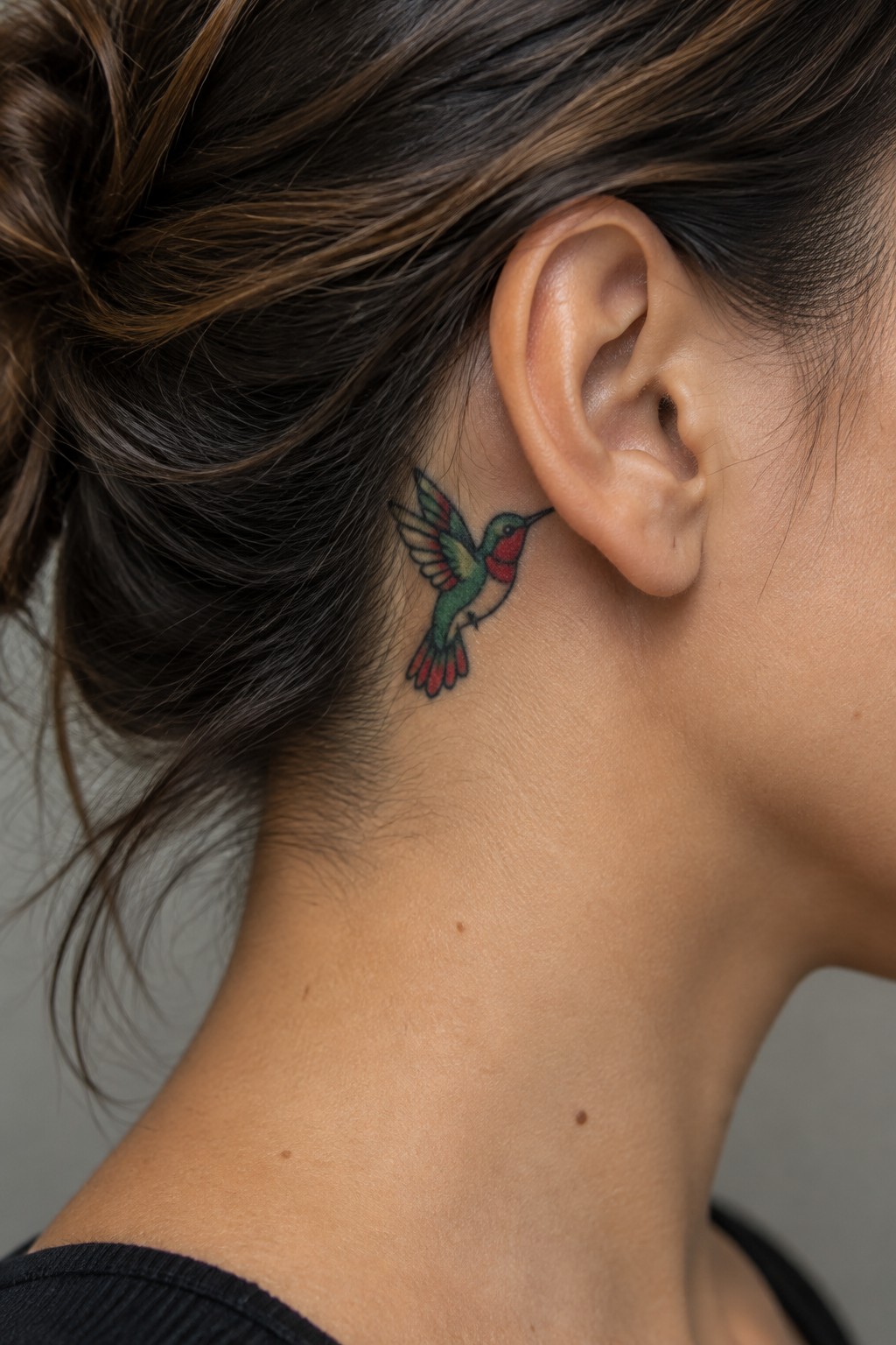 Small red and green hummingbird tattoo behind a woman’s ear, just below the hairline.