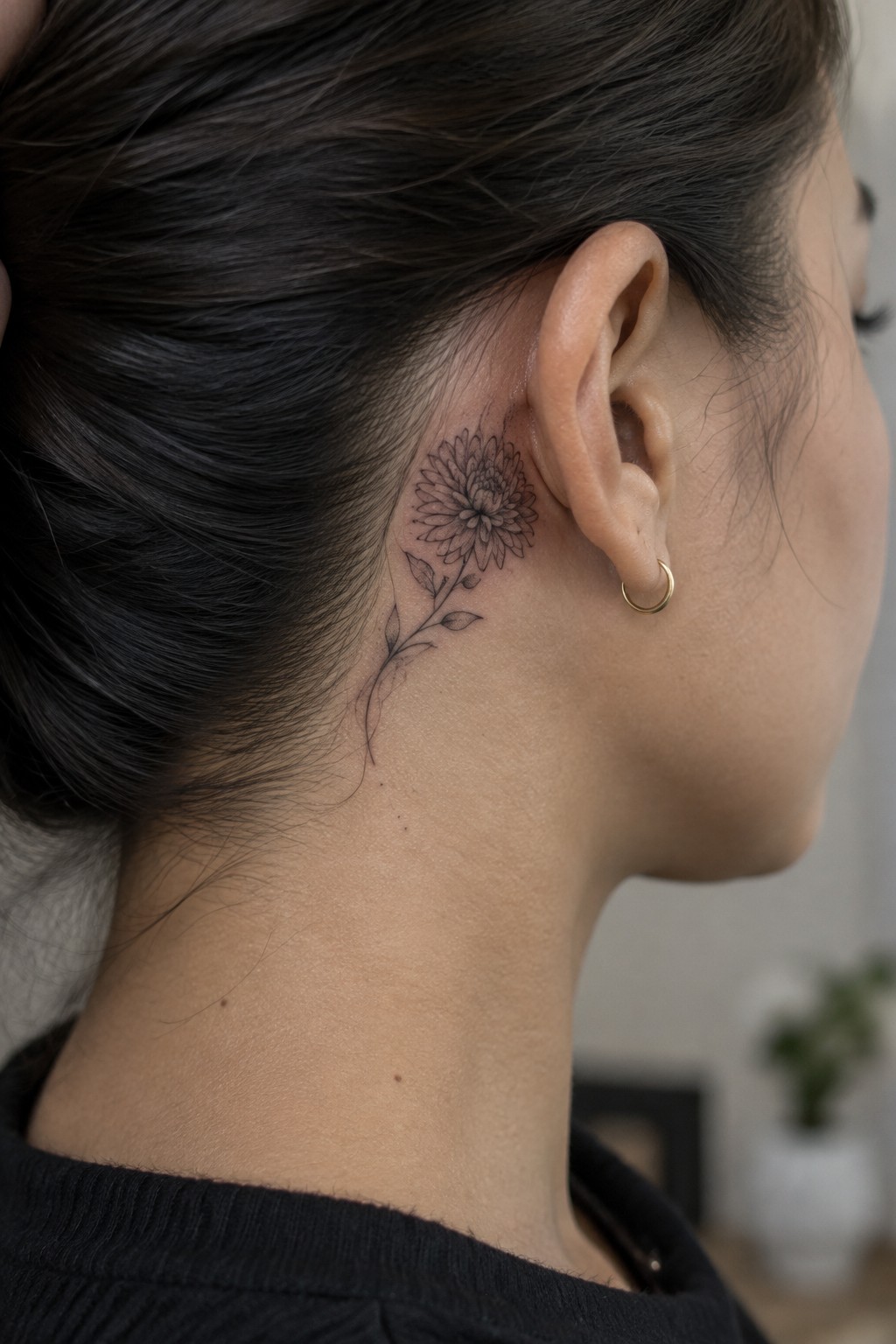 Fine-line black flower tattoo with a leafy stem behind a woman’s ear, above her neck.