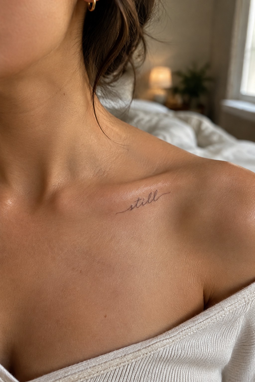 Small fine-line script tattoo reading “still” on a woman’s collarbone.