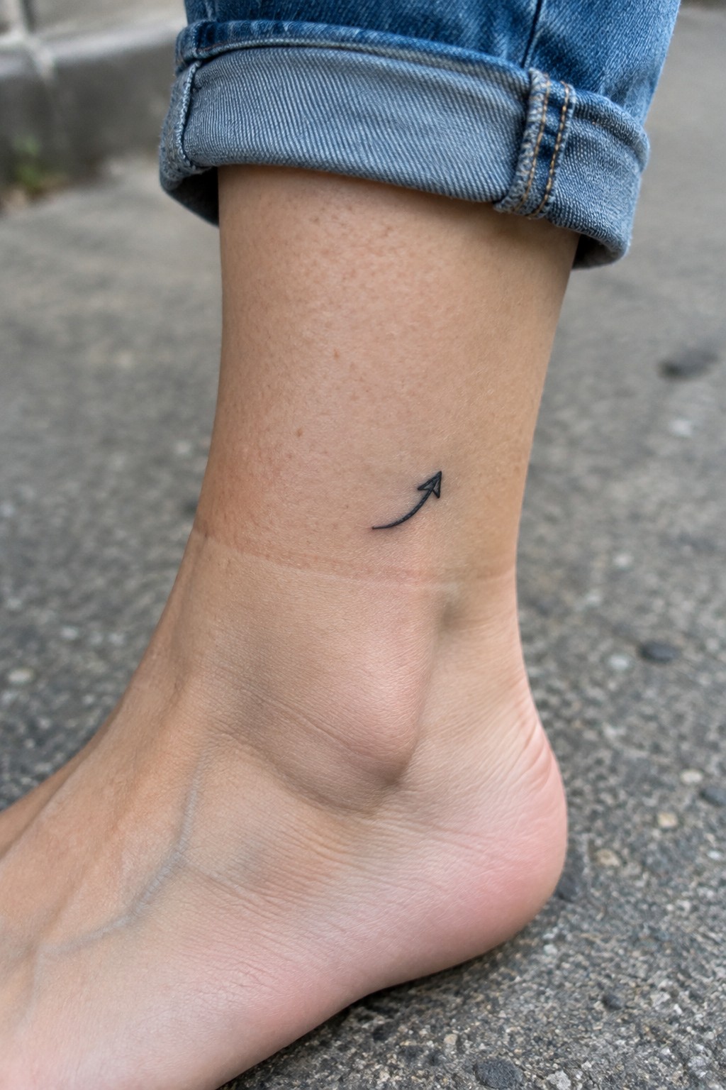 Small black curved arrow tattoo on the outer ankle above the heel.