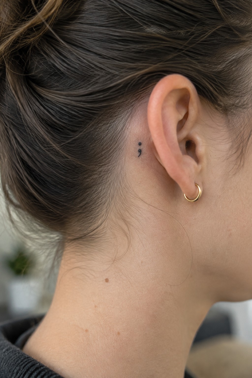 Small black semicolon tattoo behind a woman’s ear, above a gold hoop earring.