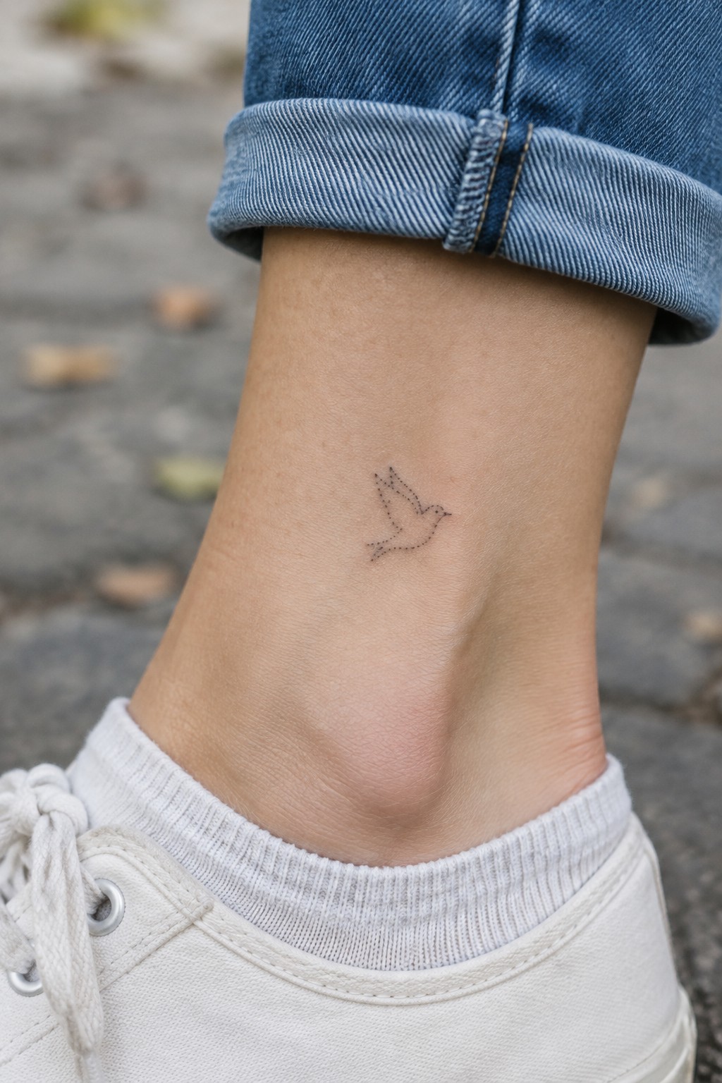 Small bird tattoo on ankle above white sneaker and cuffed blue jeans