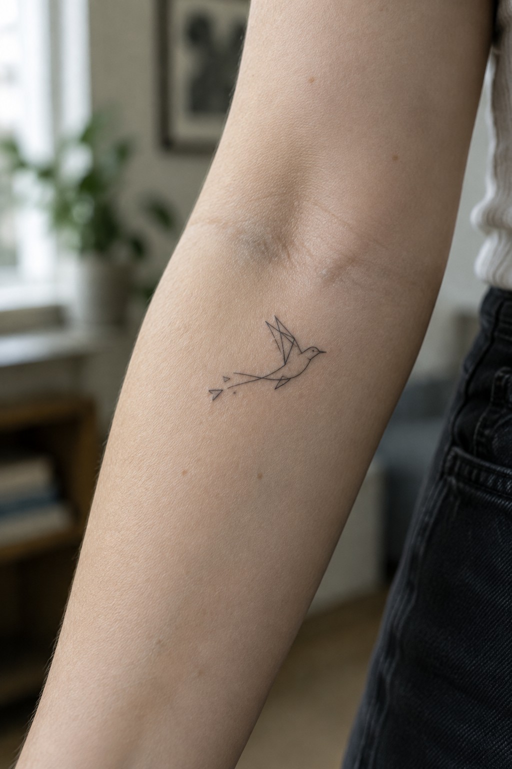 Minimalist geometric bird tattoo on light-skinned arm next to dark jeans