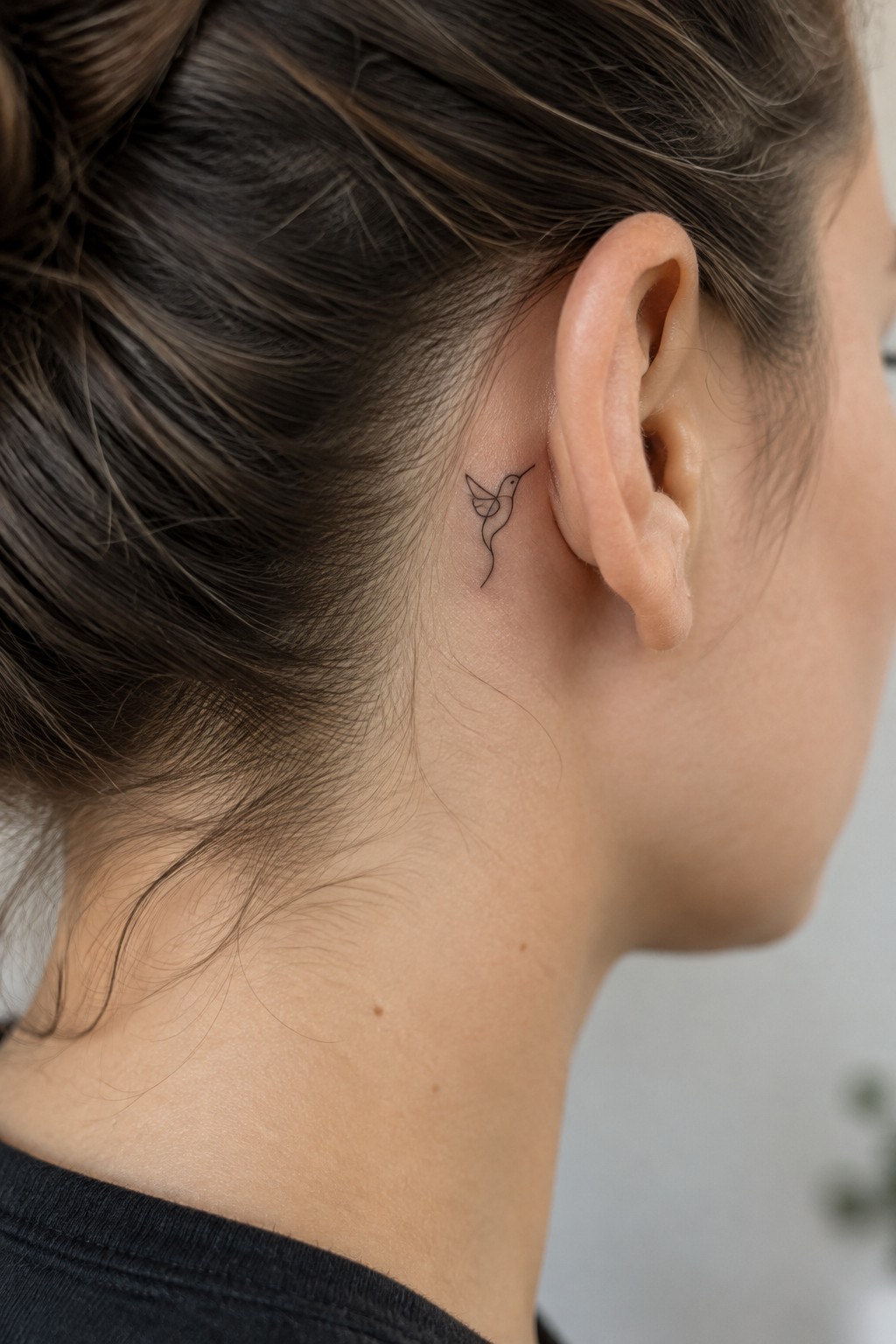 Woman's ear and neck with small hummingbird tattoo, brown hair in bun, black top