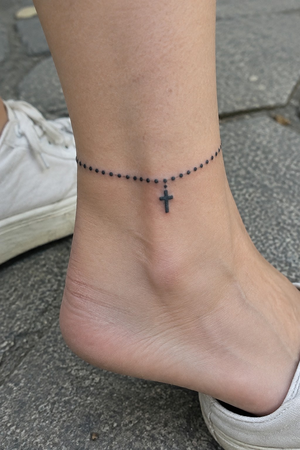 Rosary bead tattoo with cross on ankle above white sneaker on pavement